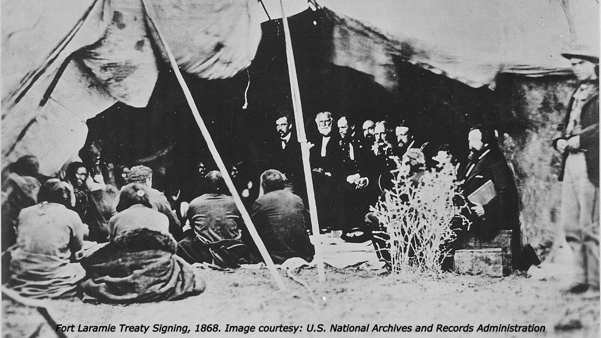 Many men with beards sit inside an open tent, with many Native Americans sitting on the ground in front of them.  