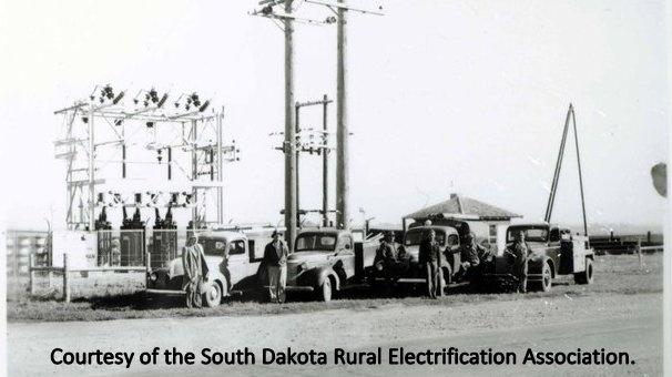 Archival photo of a electrical substation with four trucks and workers standing in front of it. 
