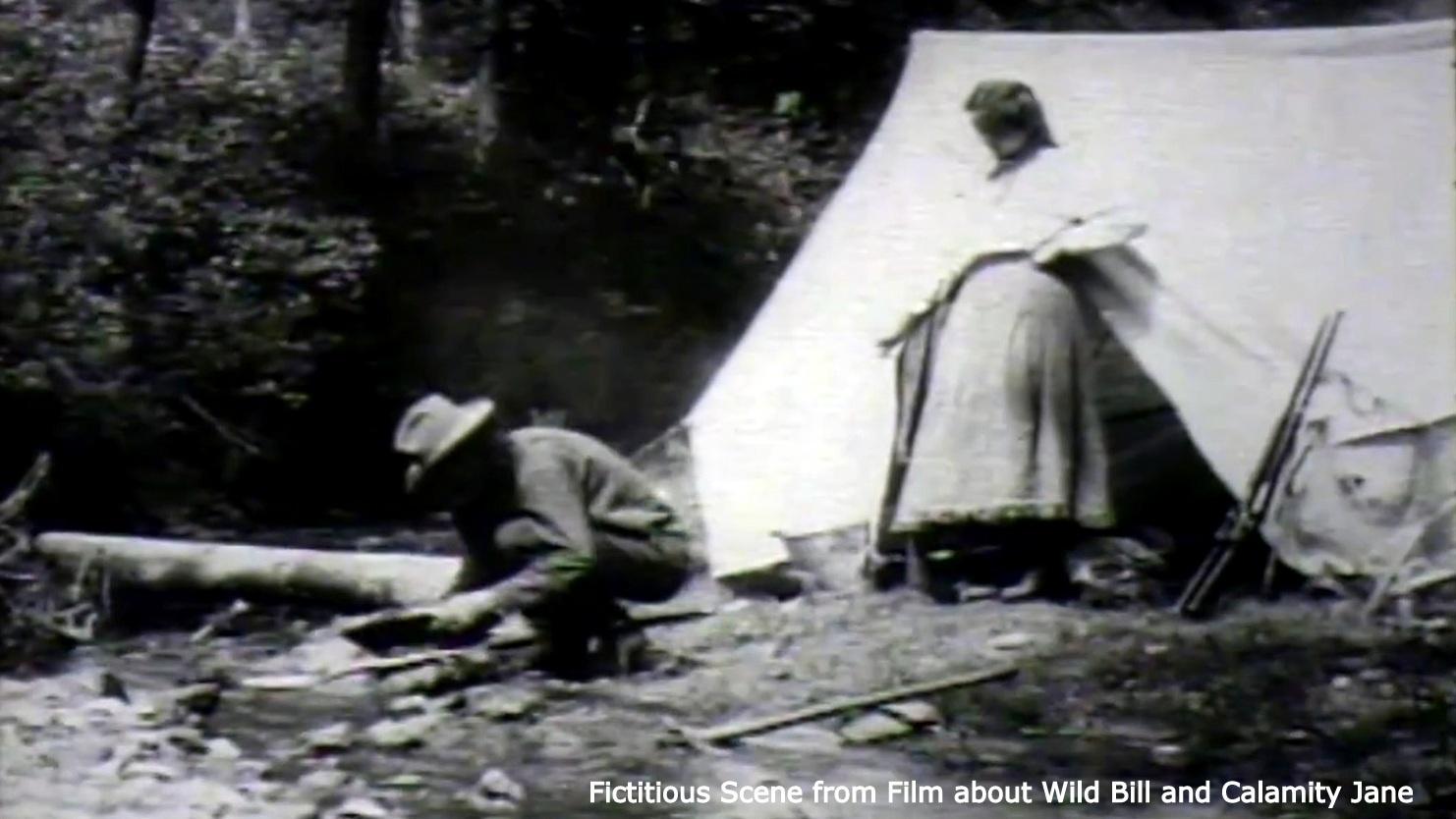 An image of a man and a woman near a tent. The man is panning for gold, and the woman stands behind him. 
