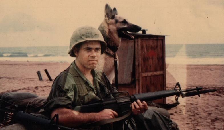 Photo of a soldier holding a machine gun with a German shepherd sitting next to him. 