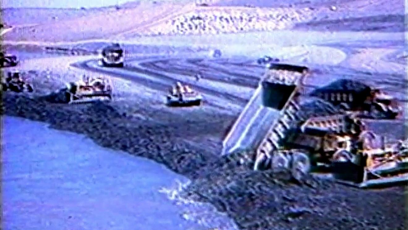 Archival photo of the building of Oahe Dam. A large dump truck is dumping dirt and rocks near the water. Four bulldozers are moving the rock and dirt to form the dam. Two additional dump trucks are shown in the background. 
