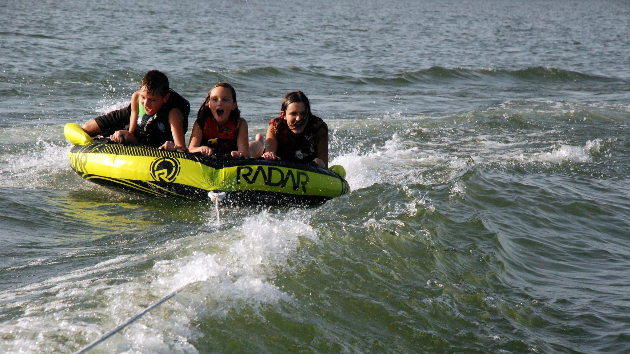 One boy and two girls are riding on a towable tube on the Missouri River.  There is a large wave in front of them and then are getting wet from water splashing on them. 