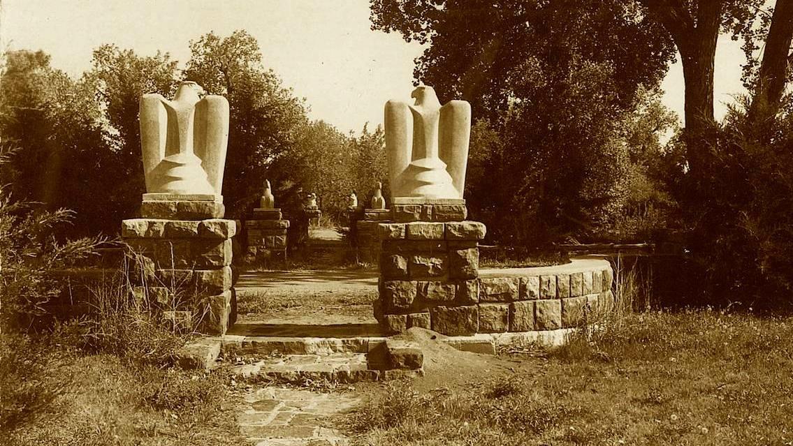 Archival photo of American Island near Chamberlain, SD. Two large bird statues are placed on either side of a walking path. There are many trees in the background.   