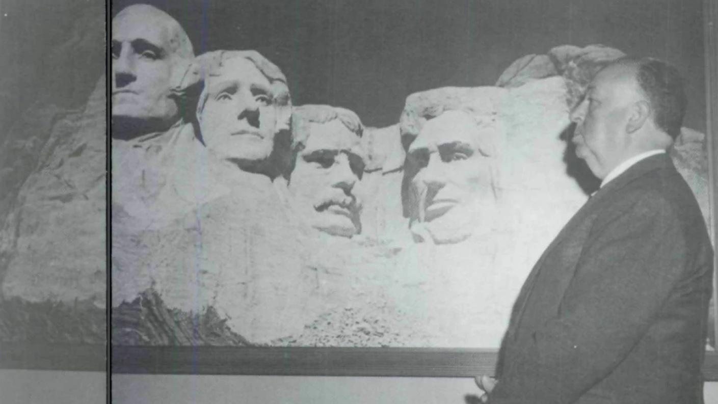 Alfred Hitchcock standing next to a photo of Mount Rushmore.