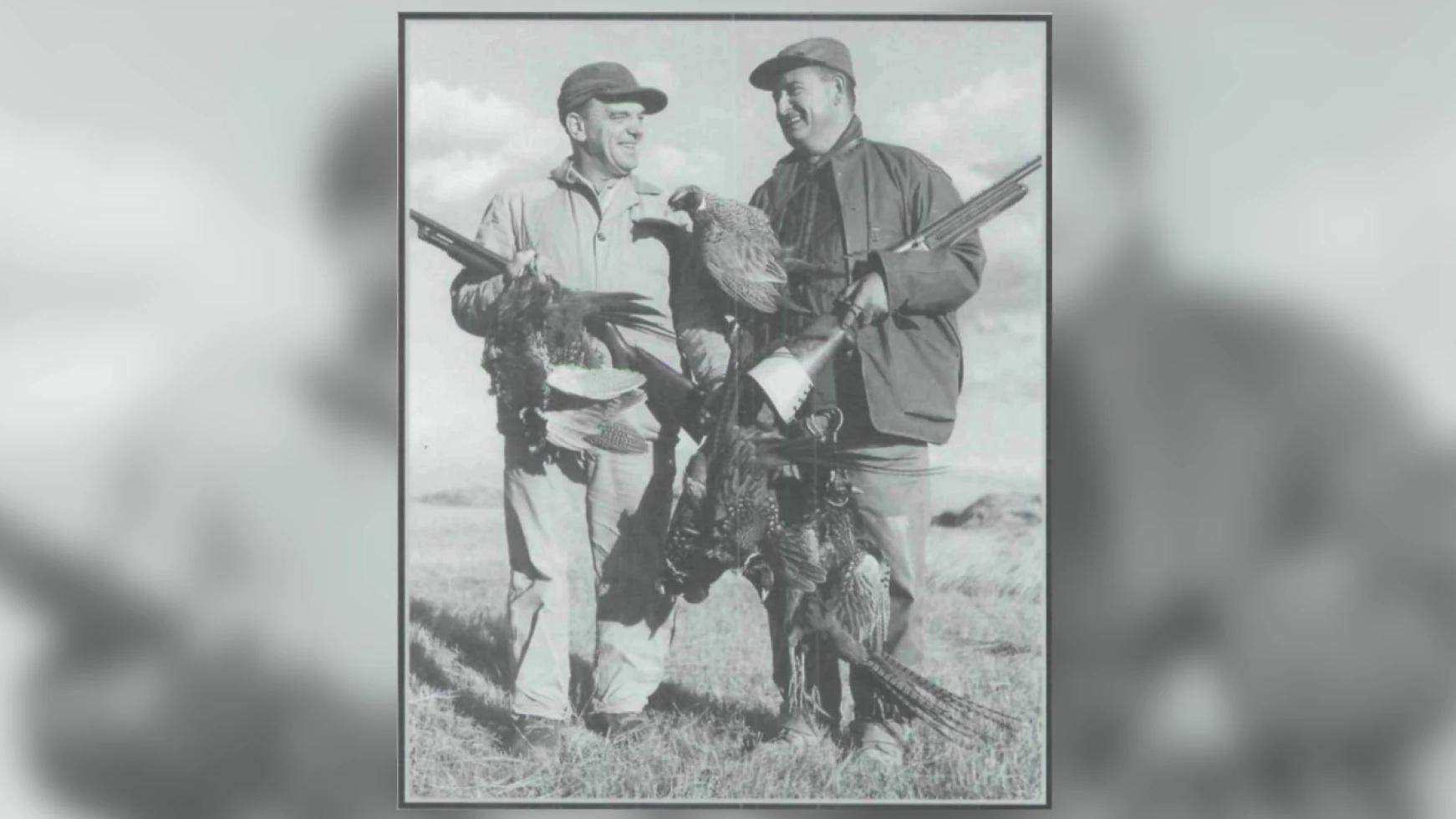 Two hunters each holding a shotgun and pheasants. 