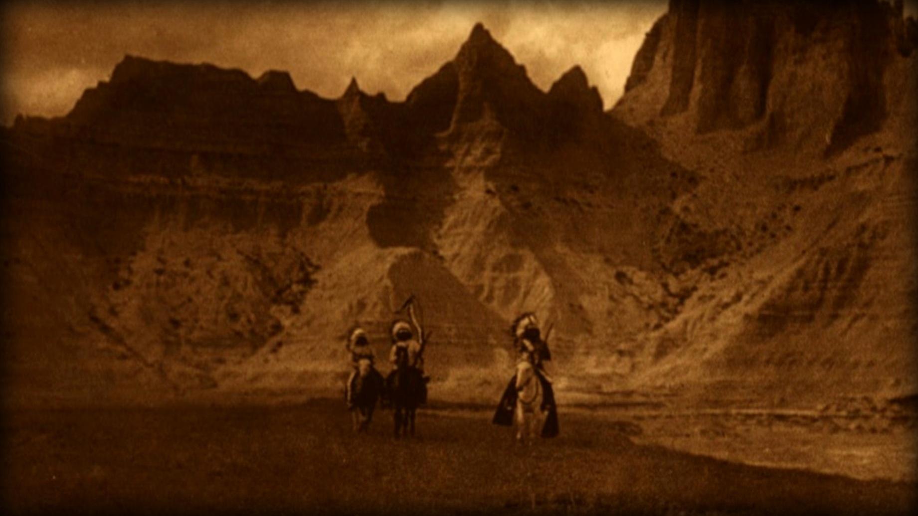 Three Native Americans with headdresses are riding horses in the Badlands. 
