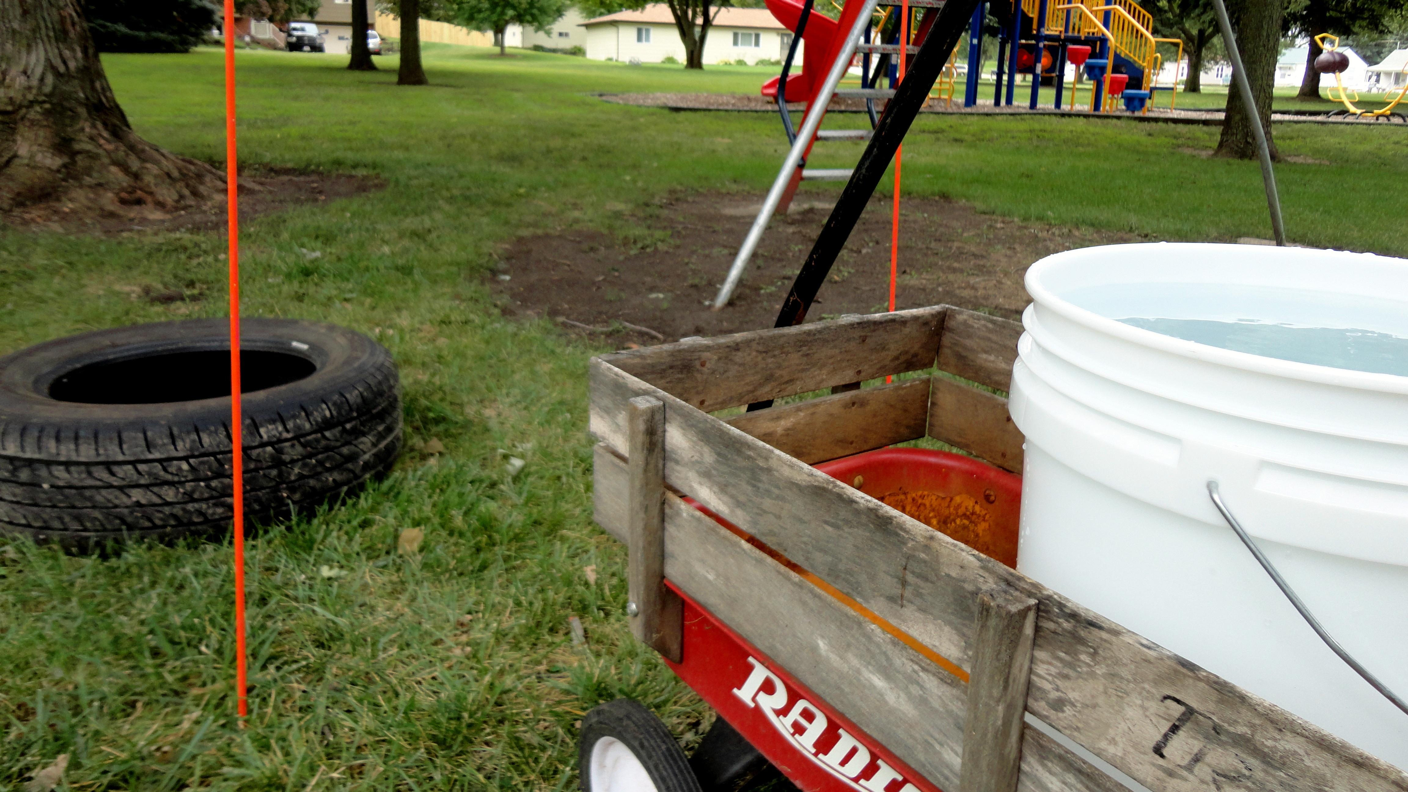 A child's red wagon is shown in a city park with a white five gallon buck in it.  