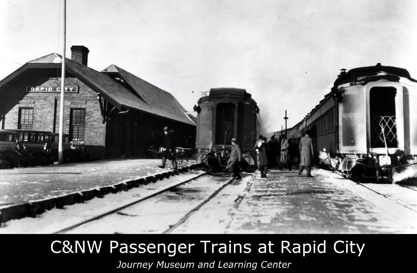 Archival photo of a train station. Two trains are shown stopped with five people walking next to them. 