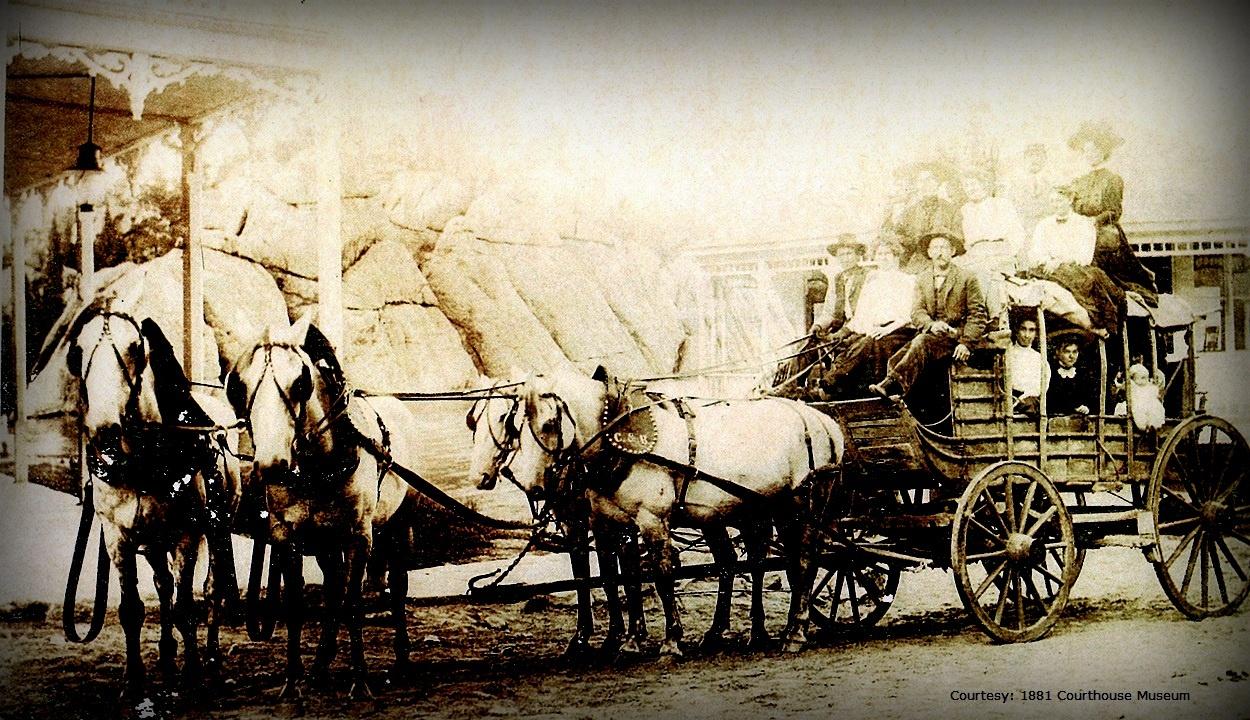 Archival image of a horse drawn stagecoach. There are four horses pulling the stagecoach. The stagecoach is full with an additional ten riders on top. 