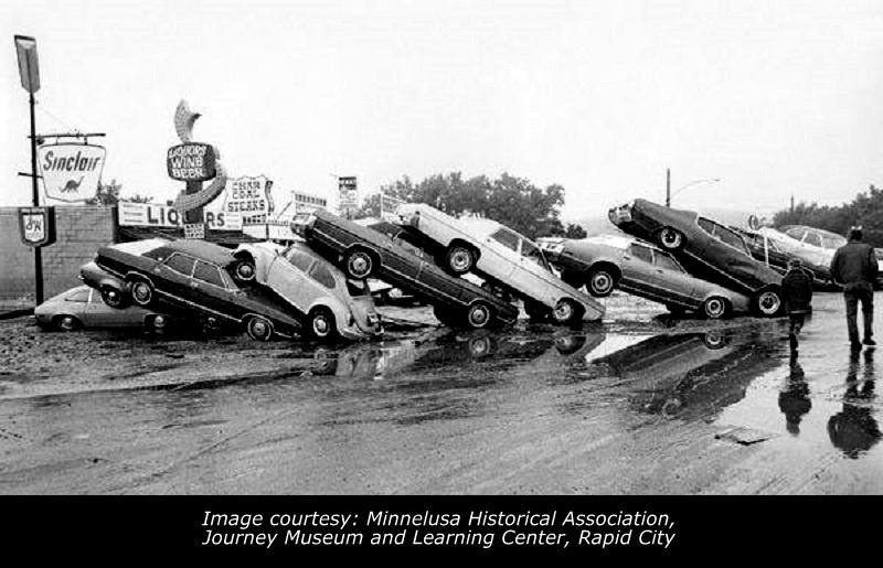 Image of Black Hills flood of 1972. Eight cars are stacked on top of each other in front of some businesses.   
