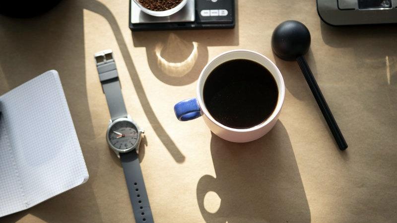 A cup of coffee is on a desk next to a watch, measuring spoon. notebook, and scale.