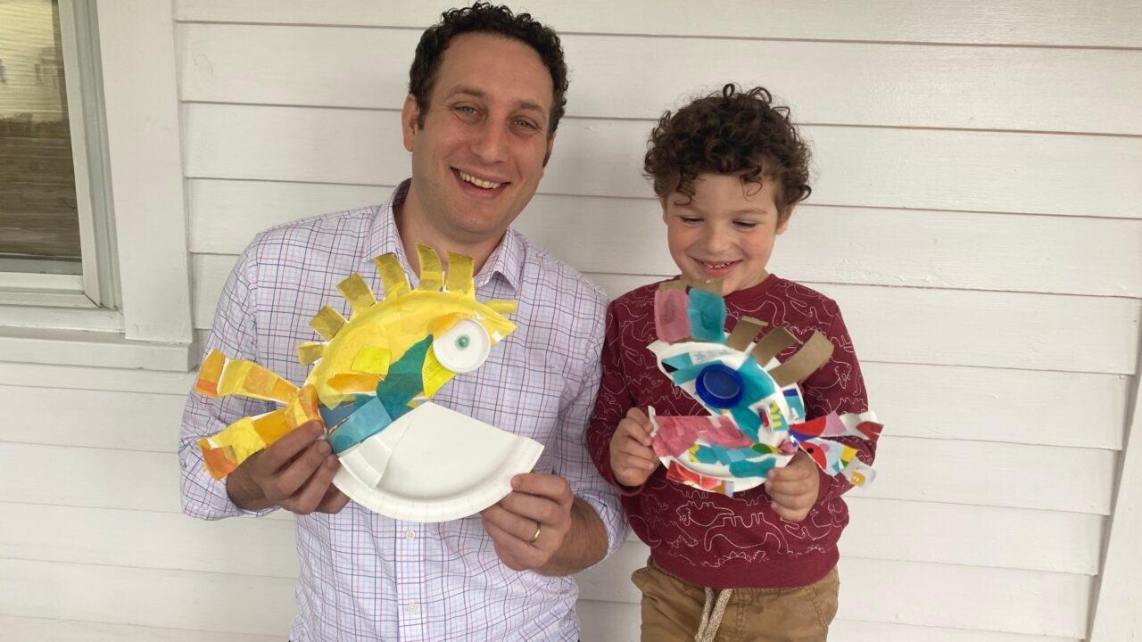 Father and son are each holding a fish craft made from a paper plate. Both are smiling. 