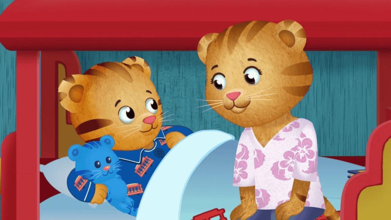 Daniel Tiger is laying in his bed covered with blankets and is holding a blue stuffed bear. His mother tiger is sitting on the bed next to him.  