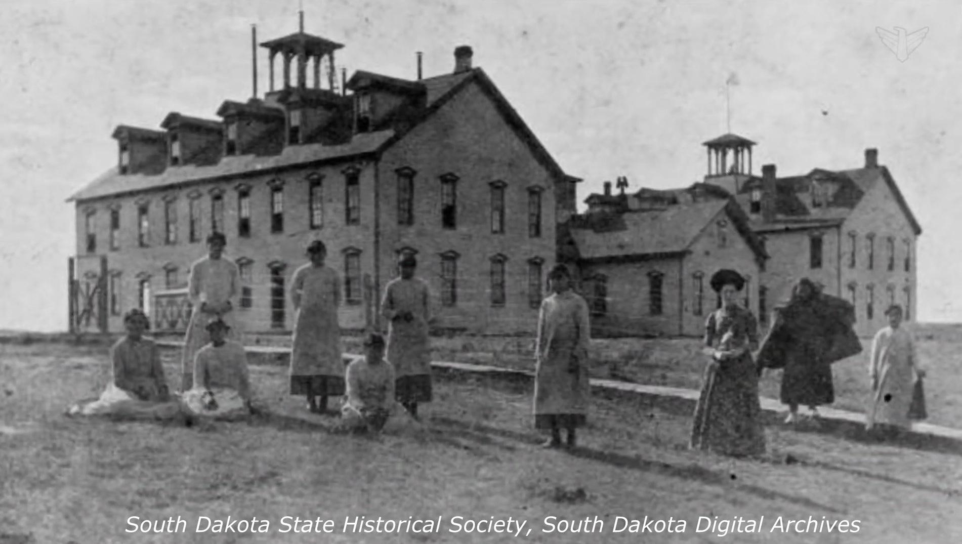 Oglala Boarding School, Pine Ridge SD - 9 students and 1 teacher are shown with three buildings in the background. Three of the students are sitting on the ground and the rest are standing. They are all wearing long dresses.   