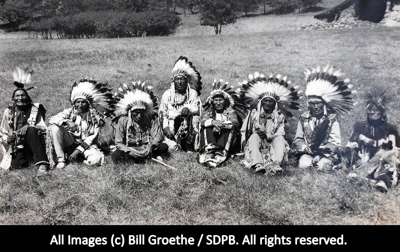 1948 photo of a reunion of the last survivors of the Battle of the Little Big Horn. Eight Native American elders, with headdresses are sitting on the grass. 