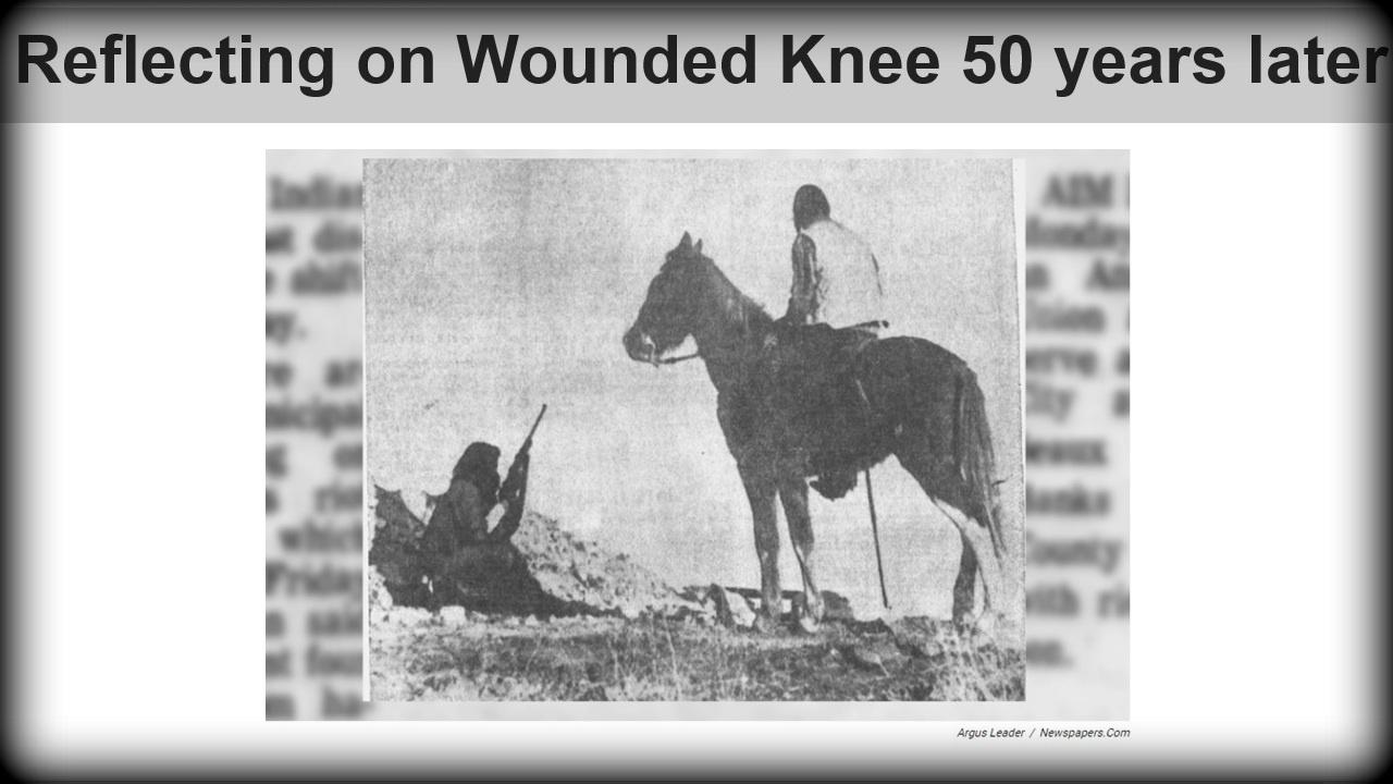 Archival photo of the 1973 protest at Wounded Knee. Profile of a Native American holding a rife is sitting on the ground, another Native American is sitting on a horse. 