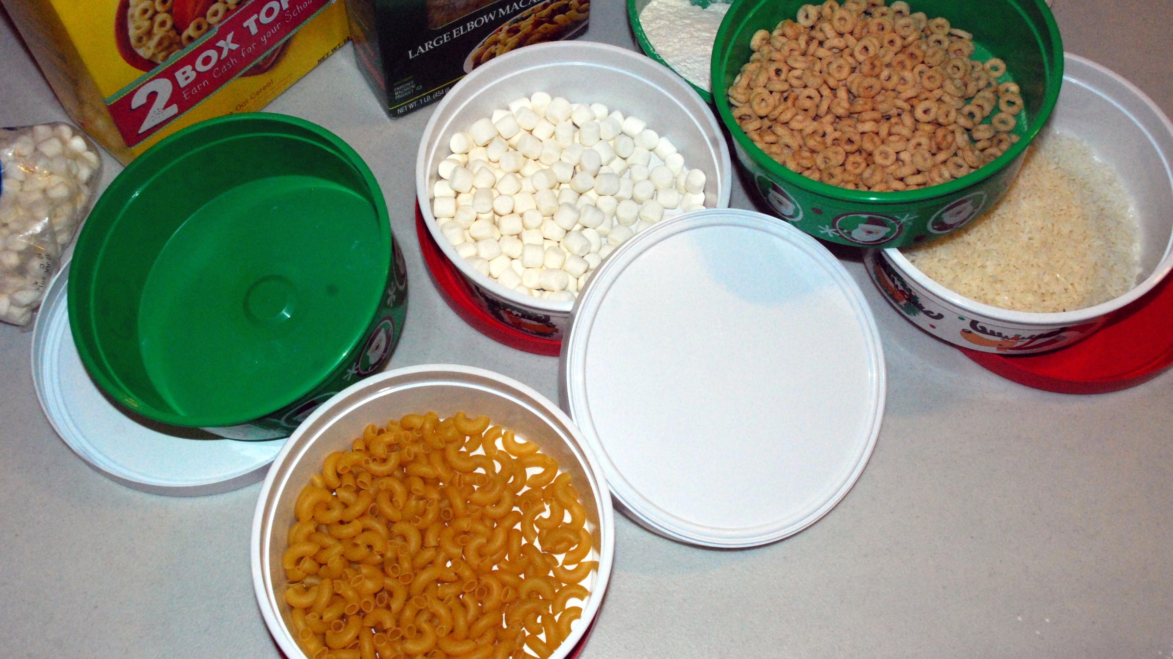 Five round storage containers. One is empty, one has elbow macaroni, the third has marshmallows in it, the forth has Cheerios, and the last one has rice.  