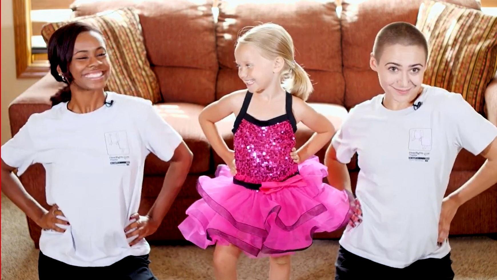 Two female dancers kneeling on either side of a little girl wearing a pink dance outfit with sequins. They are in a living room in front of a couch.