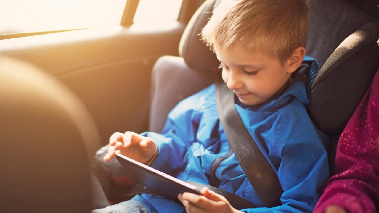 Young boy in blue coat is in his car seat playing on a tablet.