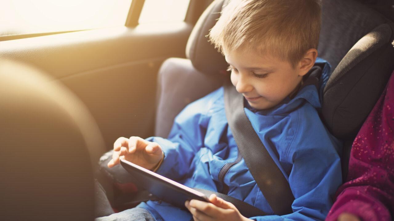 Young boy in blue coat is in his car seat playing on a tablet.