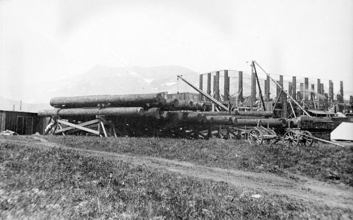 Huge logs used for construction of the Glacier Park Lodge are near a specially built siding with derricks construction materials and a freight wagon - 1911-1912