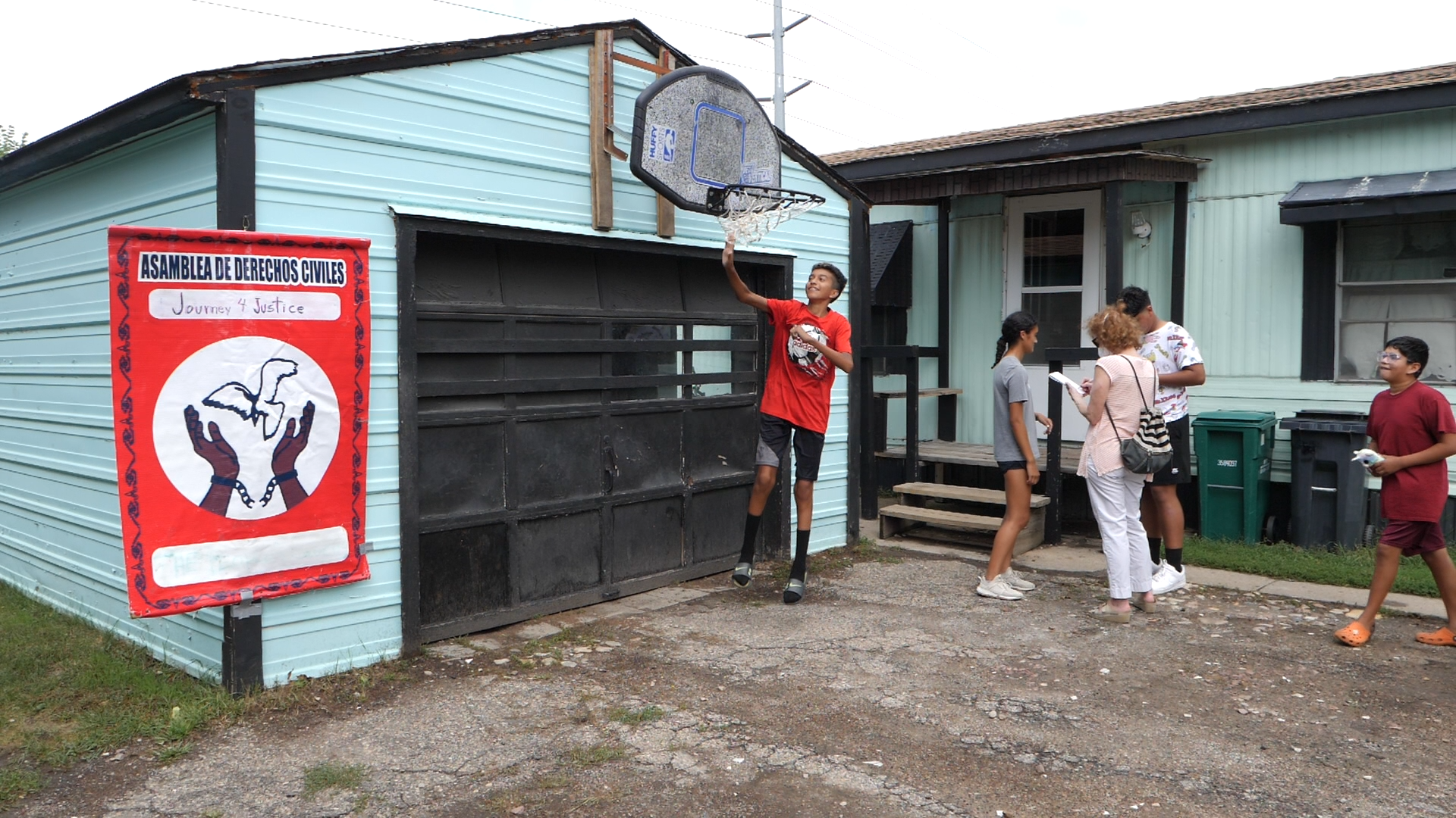 A photo of kids talking in front of a teal-colored manufactured home and one boy jumping up to hit a basketball hoop. 