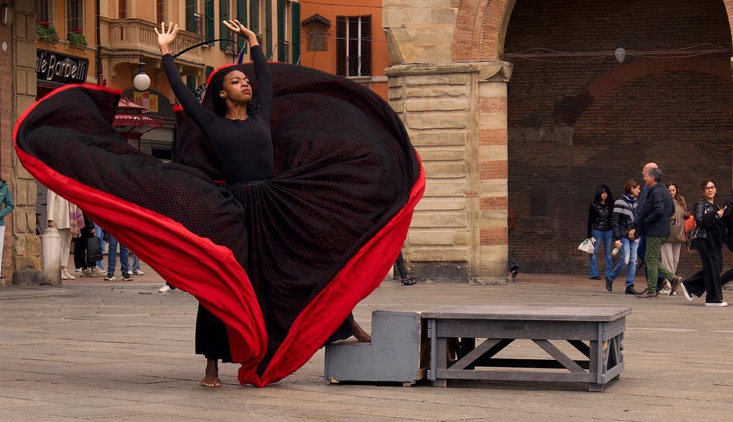 A woman stands in a busy city square. She is dancing with her arms lifted regally over her head. She has a black skirt with a red underlining that flares out around her in movement.