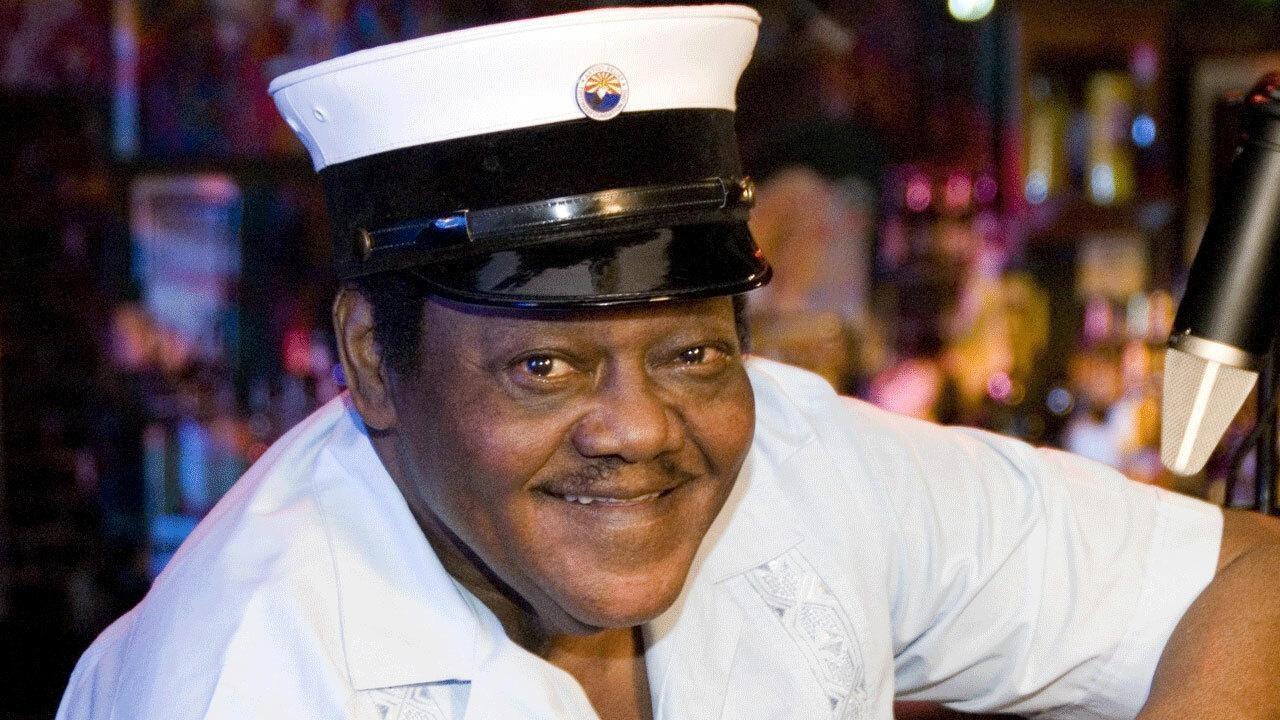 Iconic singer Fats Domino sits at a bar smiling to the camera. He wears a white and black cap and a white short sleeved shirt.