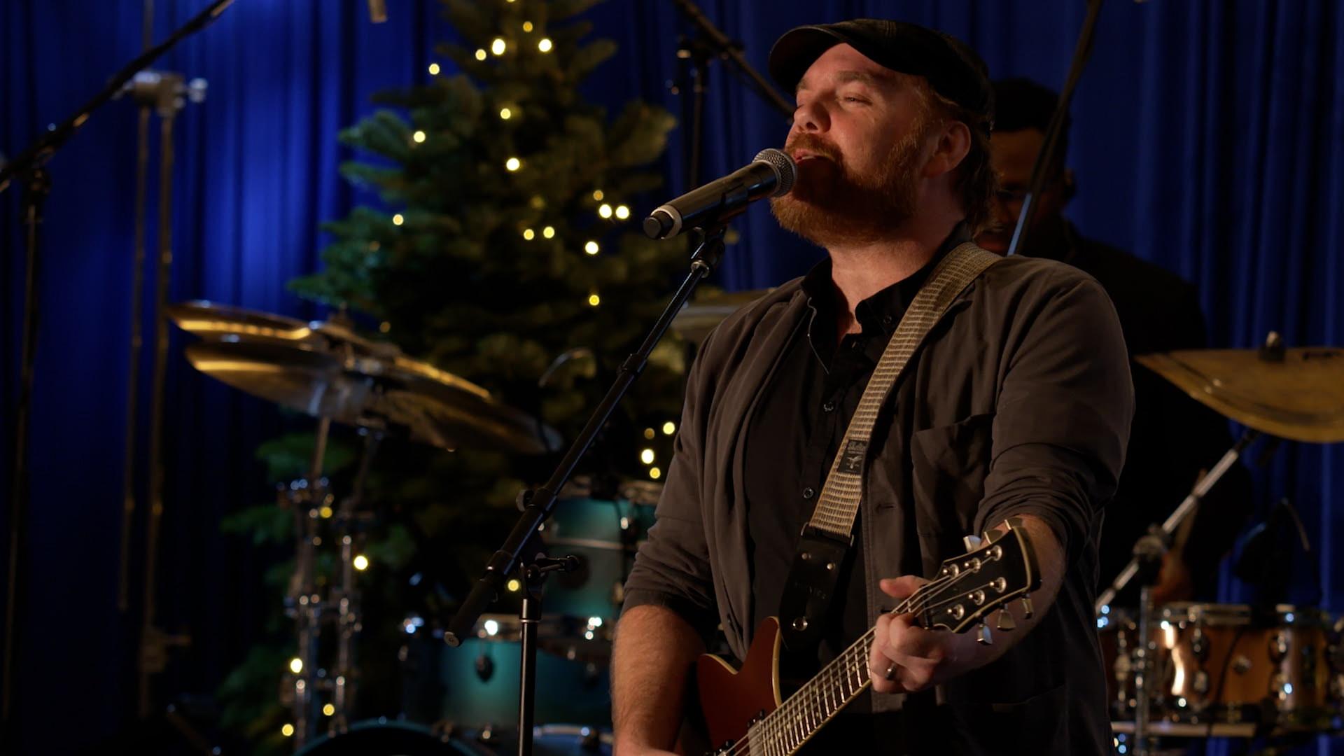 Louisiana singer songwriter Marc Broussard sings into a microphone while playing guitar. The stage is decorated with a Christmas tree.
