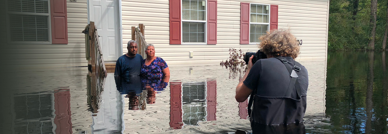 Gideon Mendel photographs a husband and wife. All three are waist-deep in water, and stand in front of a flooded house, after Hurricane Florence hit the Carolinas