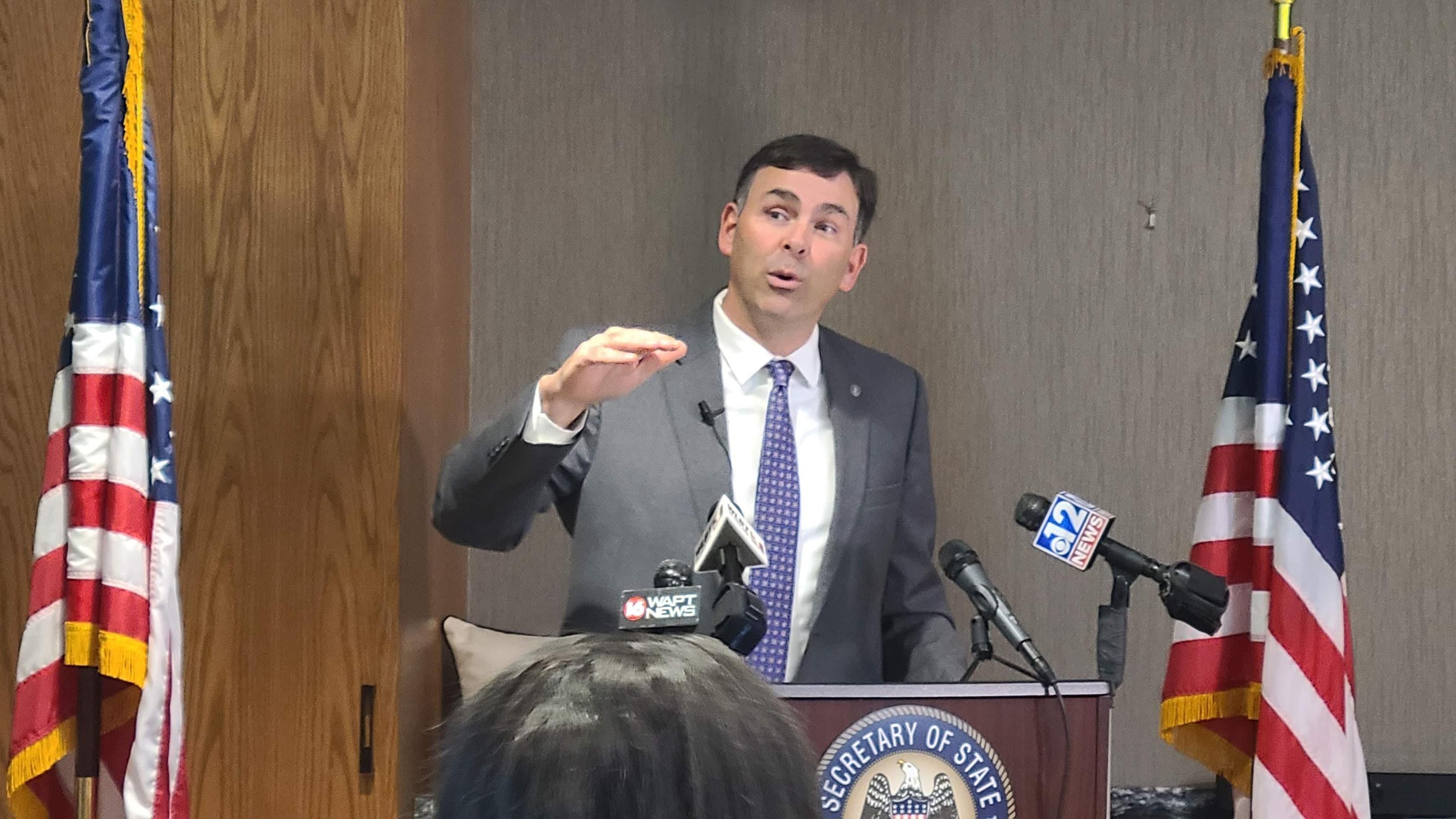Secretary of State Michael Watson stands at a podium flanked by two American Flags. He gestures with his hands as he discusses the election