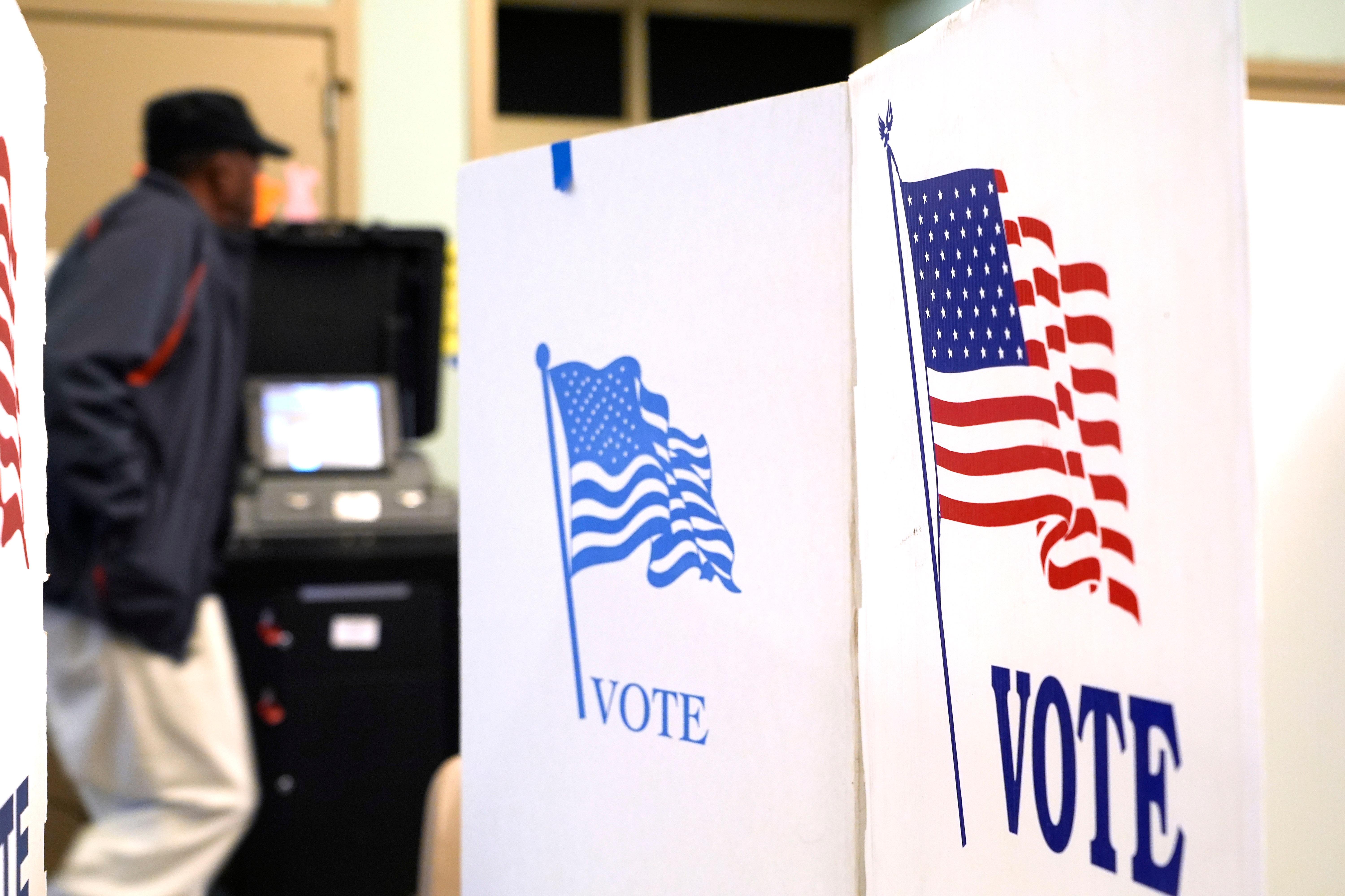 A man stands in front of a voting machine as three voter privacy stand behind him. 