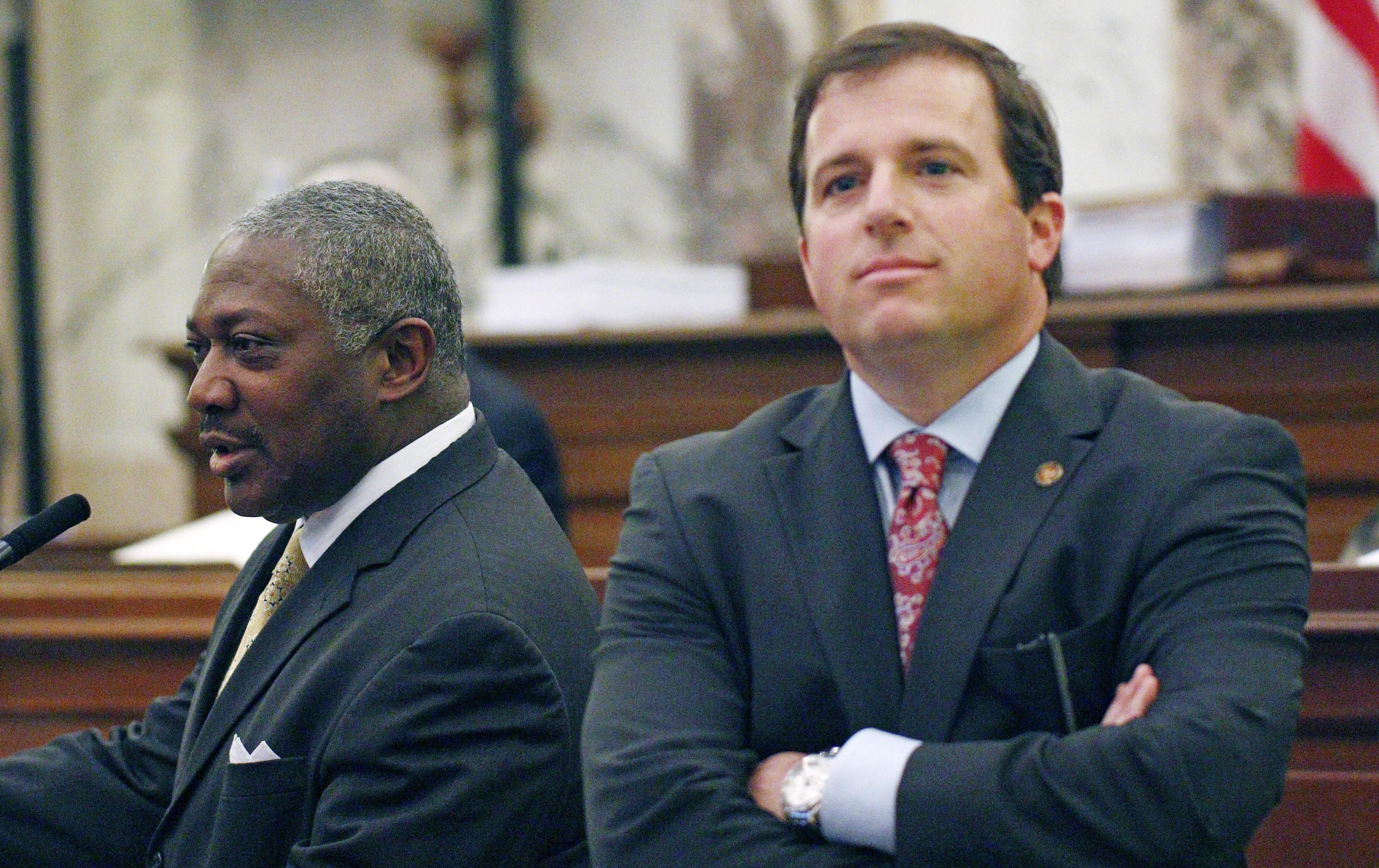 One man faces forward, listening, and another stands to the side as he speaks before a committee.