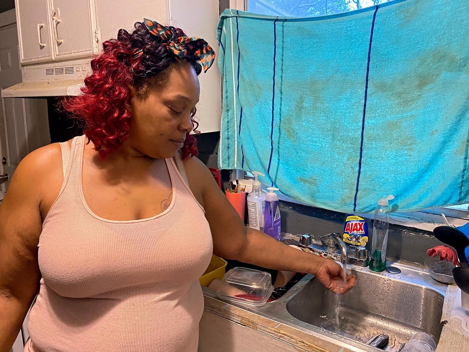 A woman stands to the side of her sink and runs water over her hand from the tap.