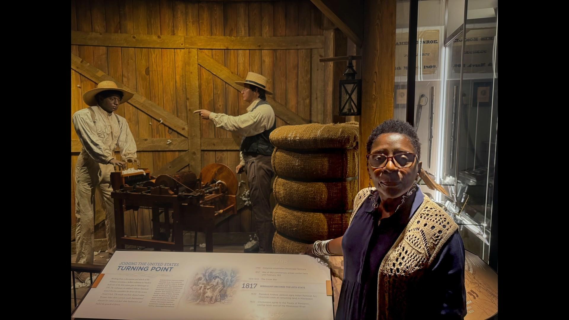 A woman stands in front of an exhibit showing an enslaved man working on a cotton gin with an overseer looking on.