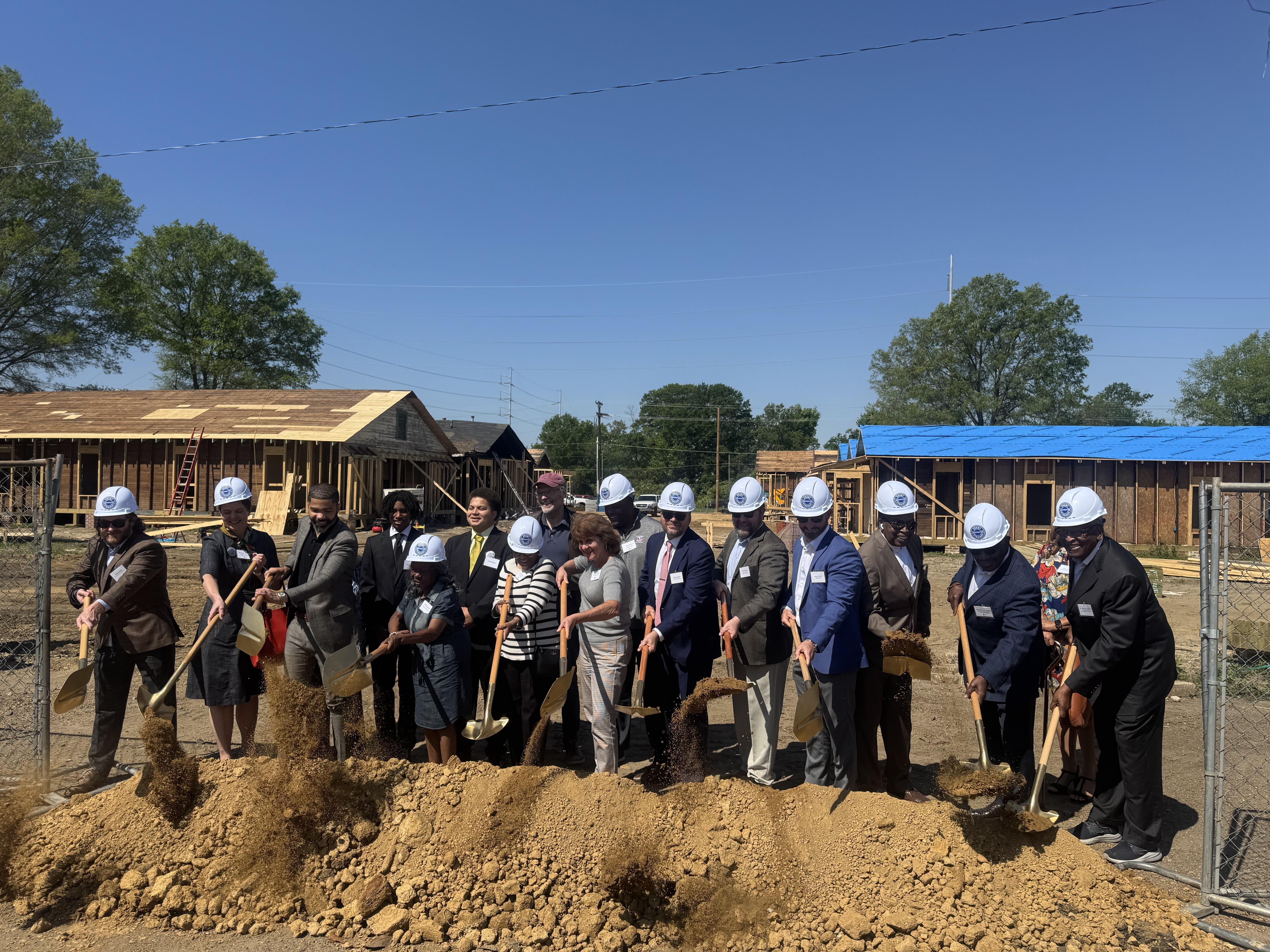 A group of officials wearing white hard hats drop dirt to the ground using golden shovels.