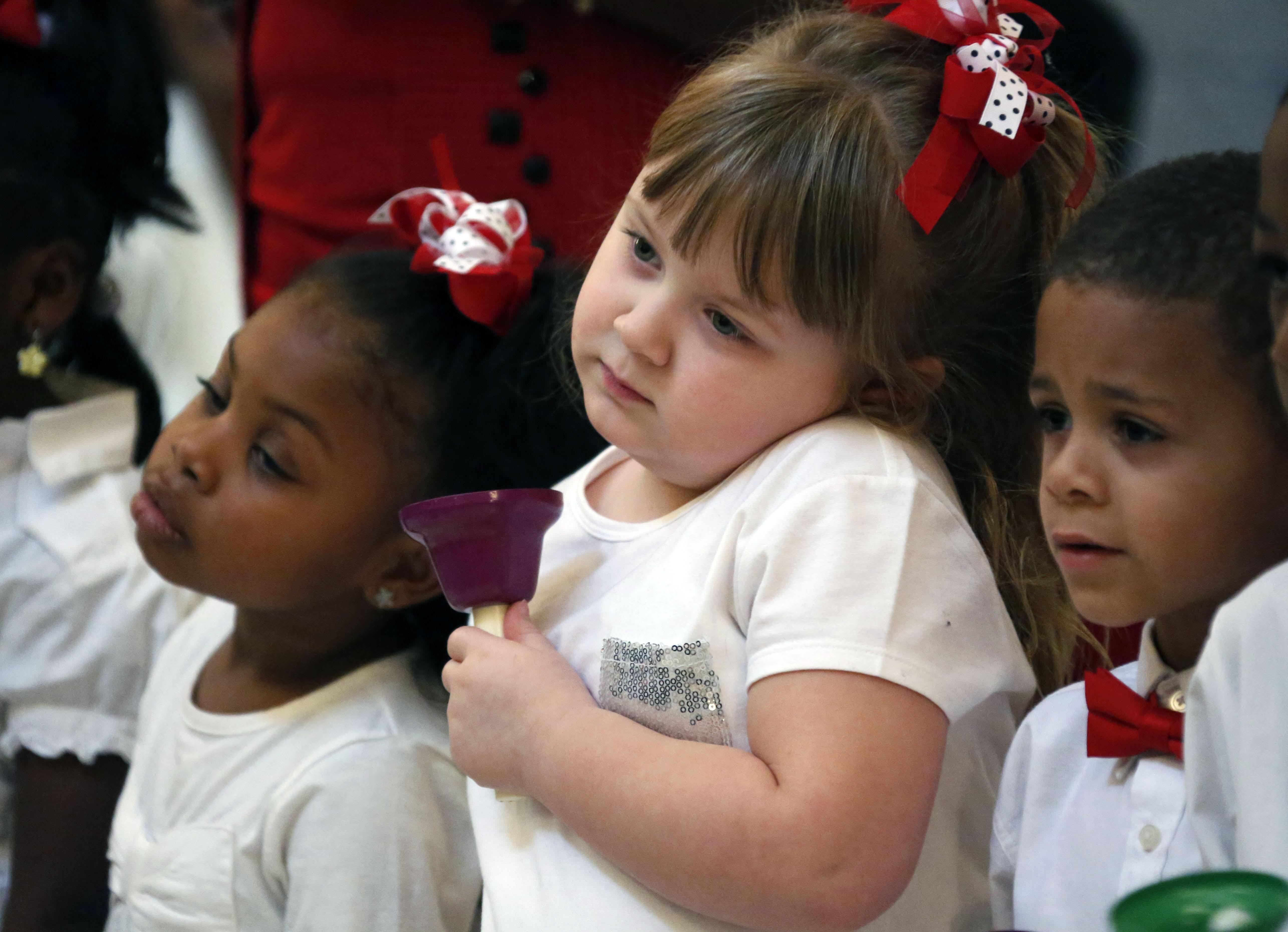 Three small children wearing white shirts stand in a row, hold different colored bells.