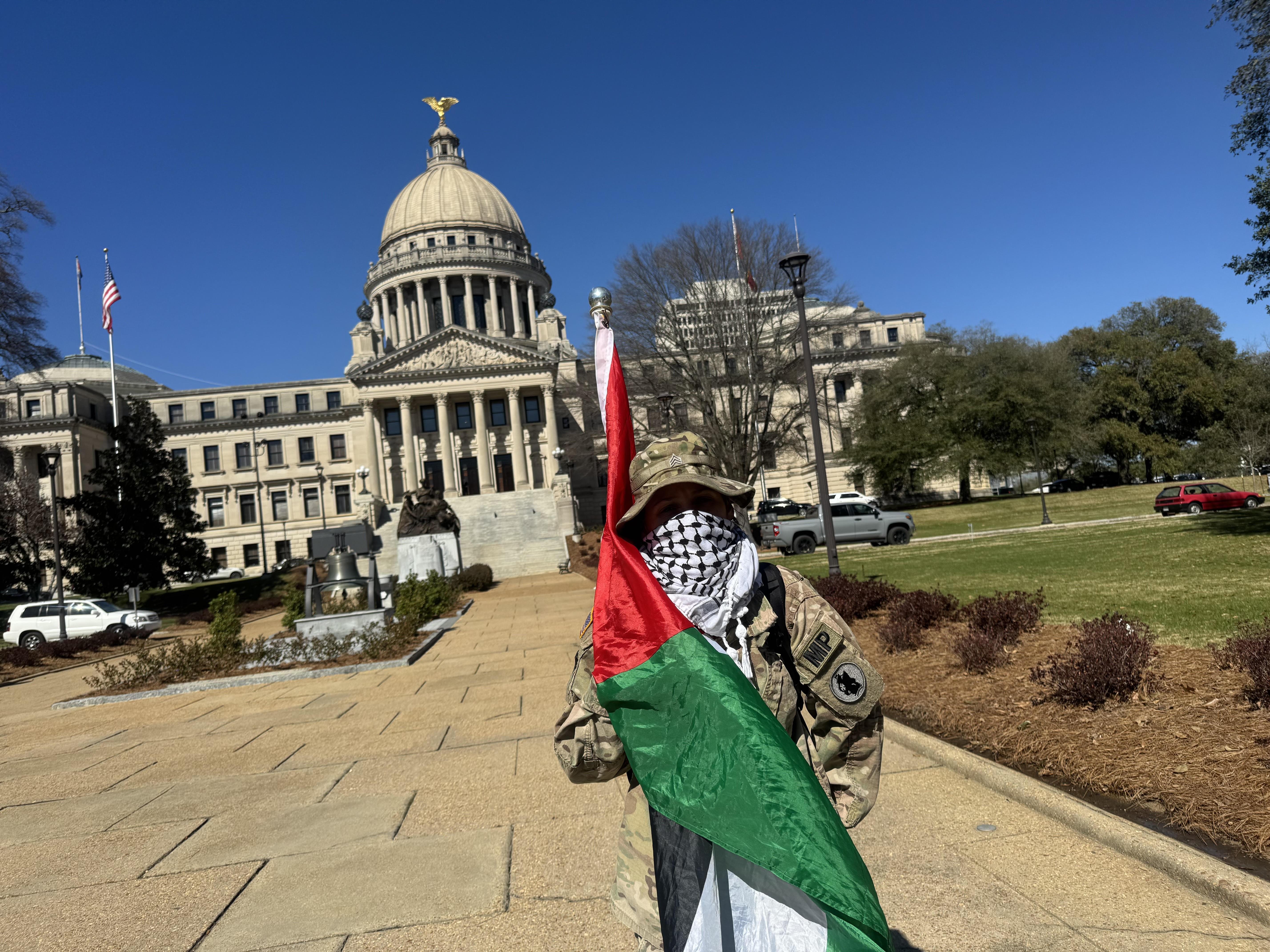 A young woman wearing a military uniform and a Keffiyeh scarf, holds a Palestinian flag.  