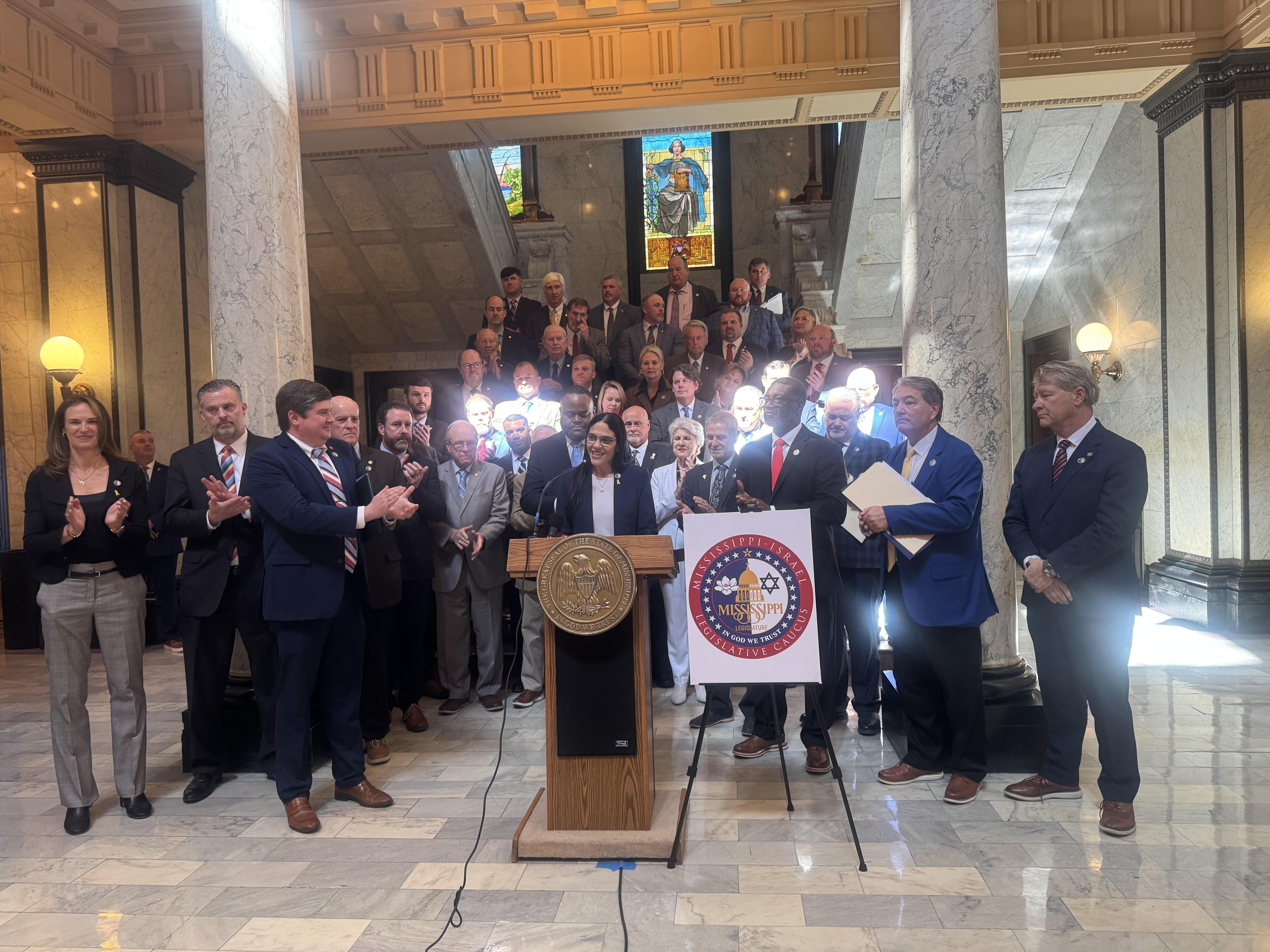 A woman stands behind a podium as a group of lawmakers stands behind her.