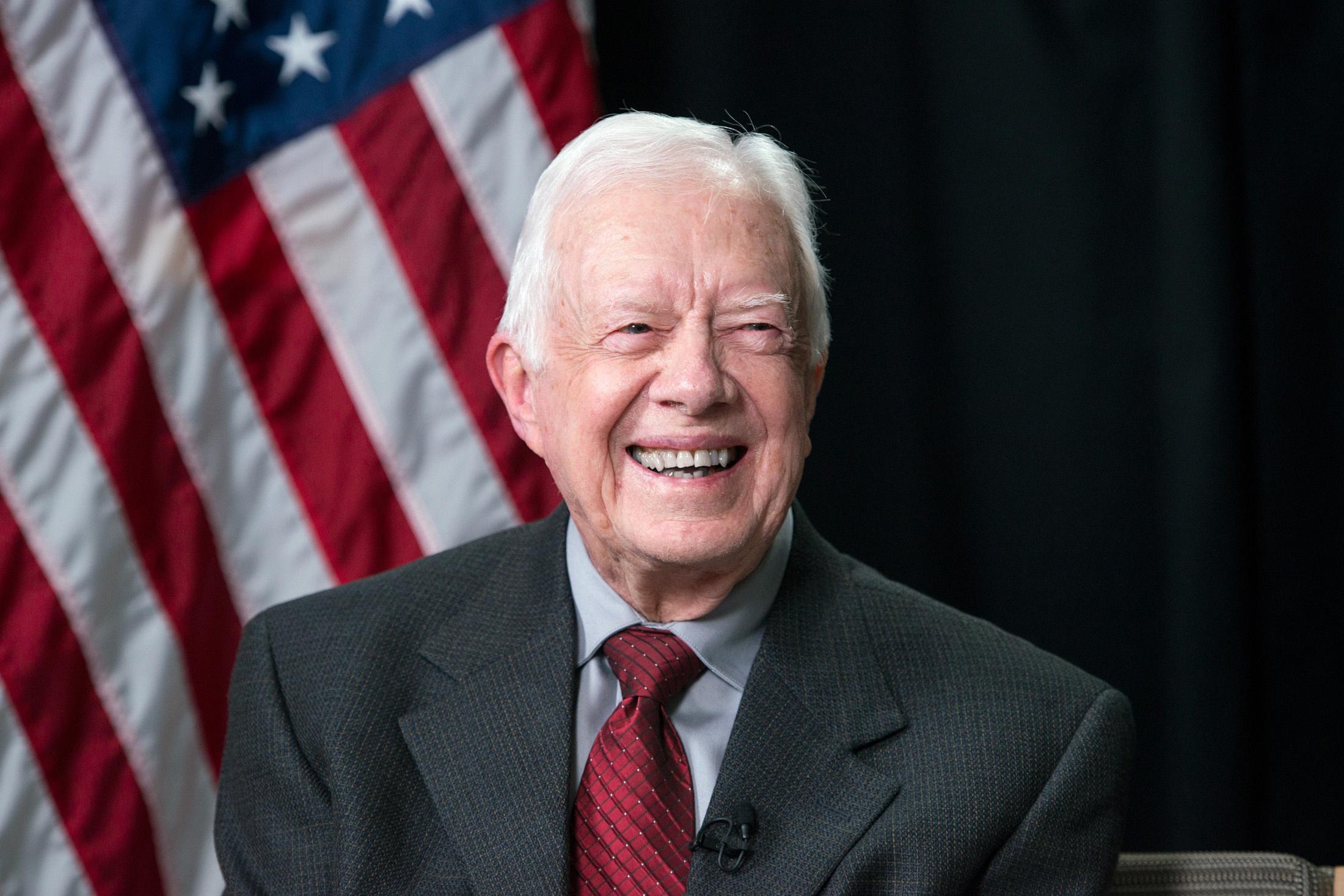 A man sits while wearing a grey suit and smiles.