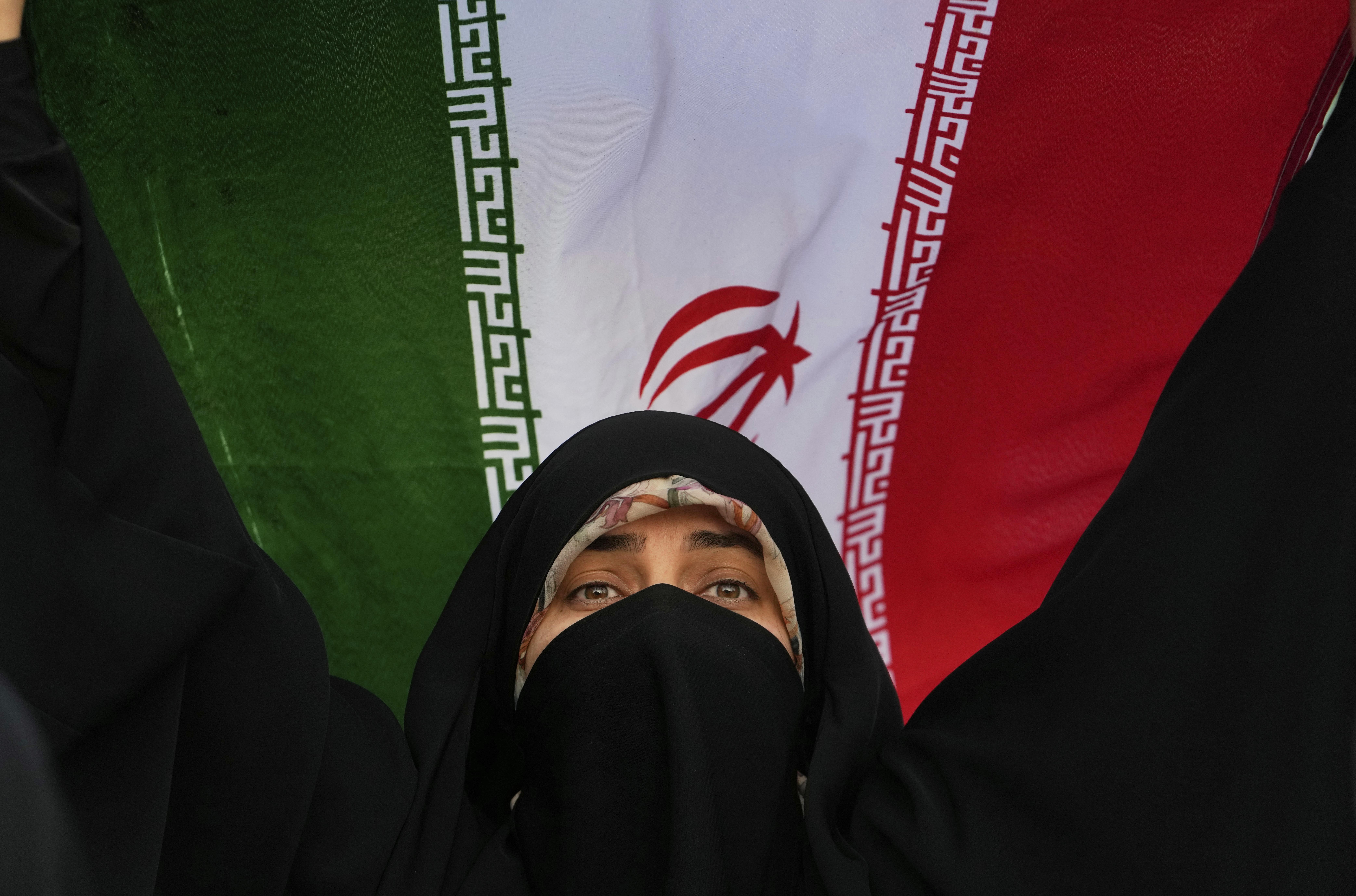 An Iranian woman wearing a black niqab holds up an Iranian flag behind her.