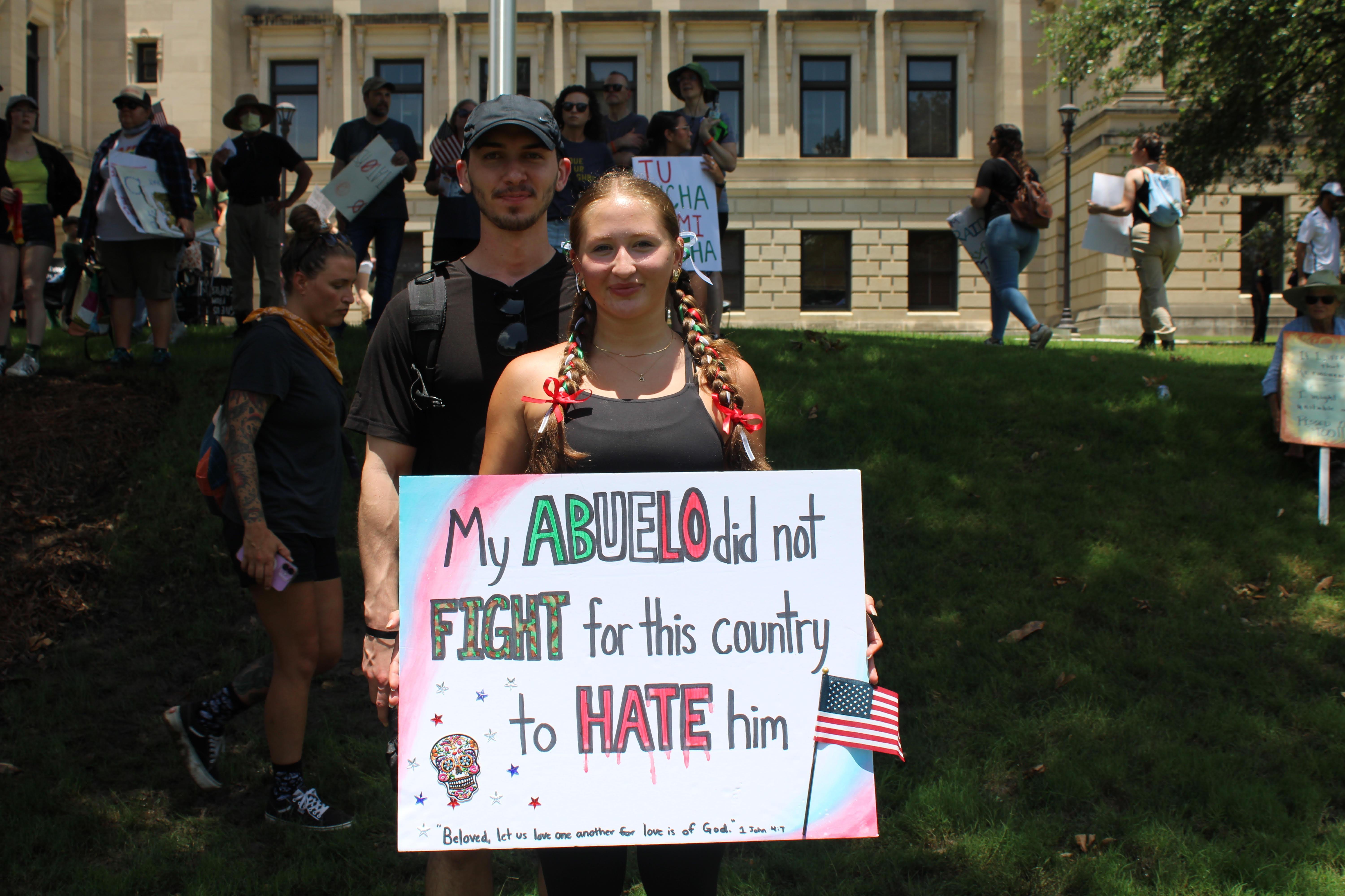 A young woman wearing the colors of the Mexican flag holds a sign reading 'my abuelo did not fight for this country to hate him.'