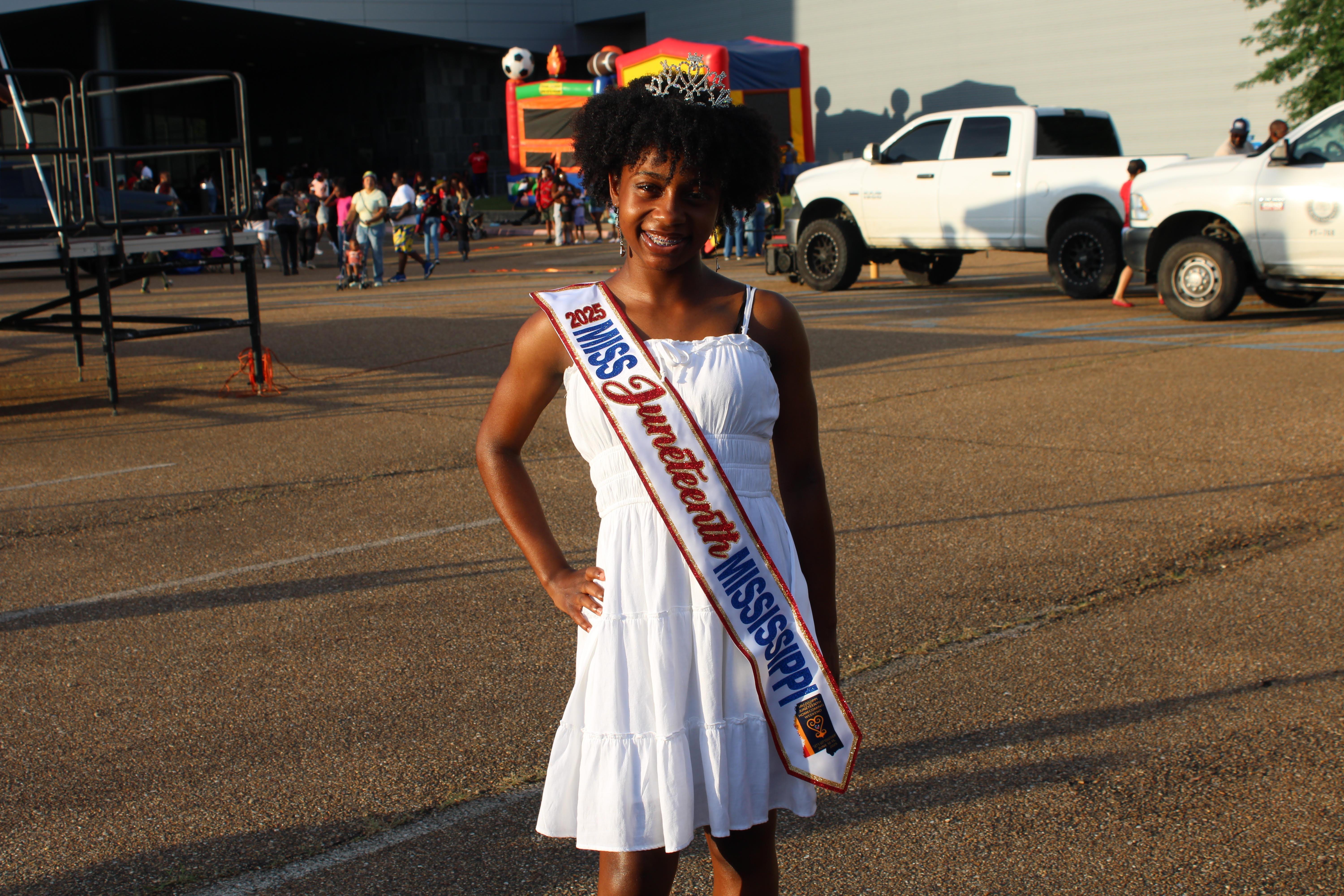 A young girl with natural hair wears a crown and sash reading '2025 Miss Juneteenth Mississippi.'