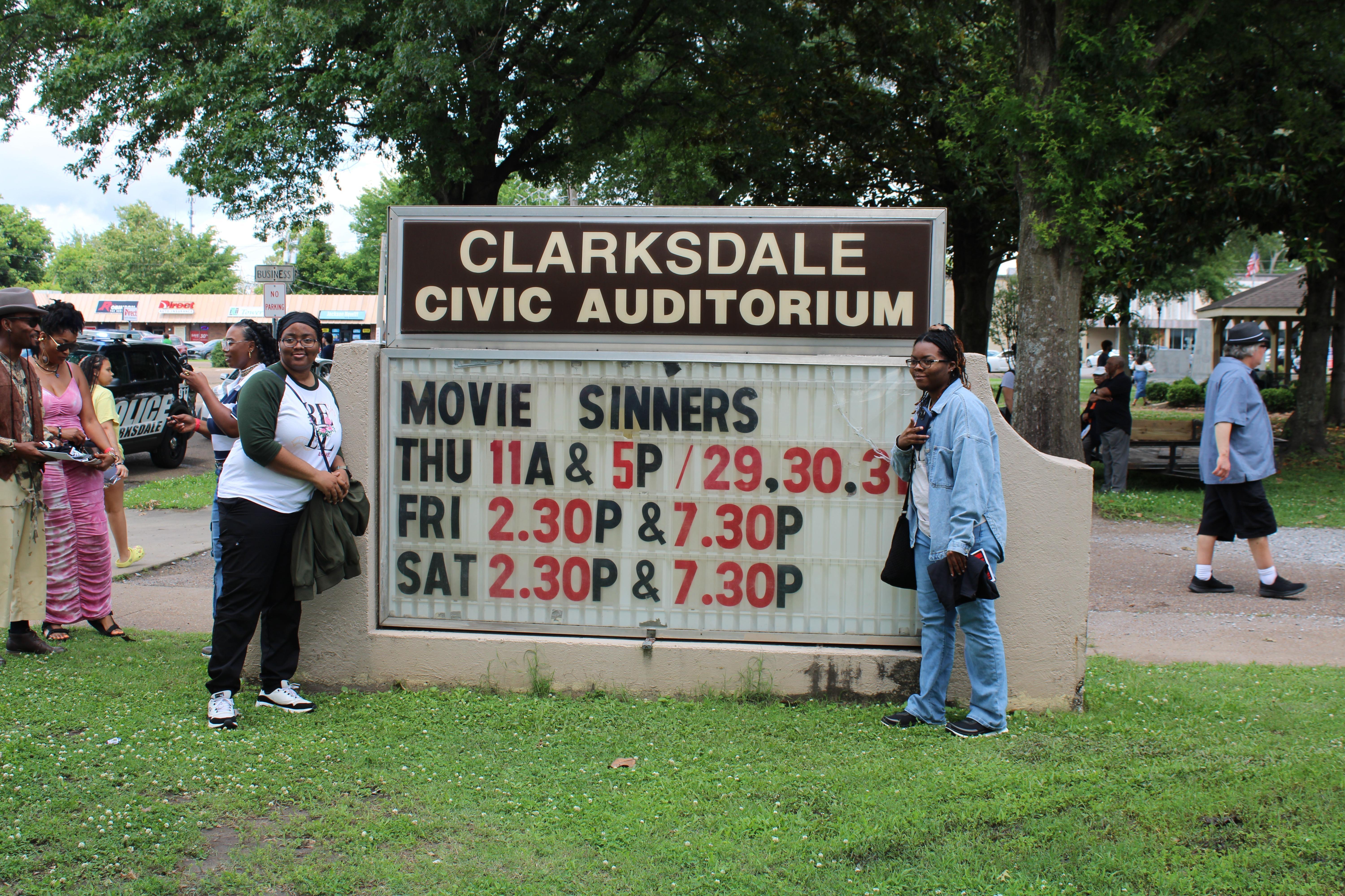 Two young women stand on either side of a marquee featuring the showtimes of the movie 'Sinners'
