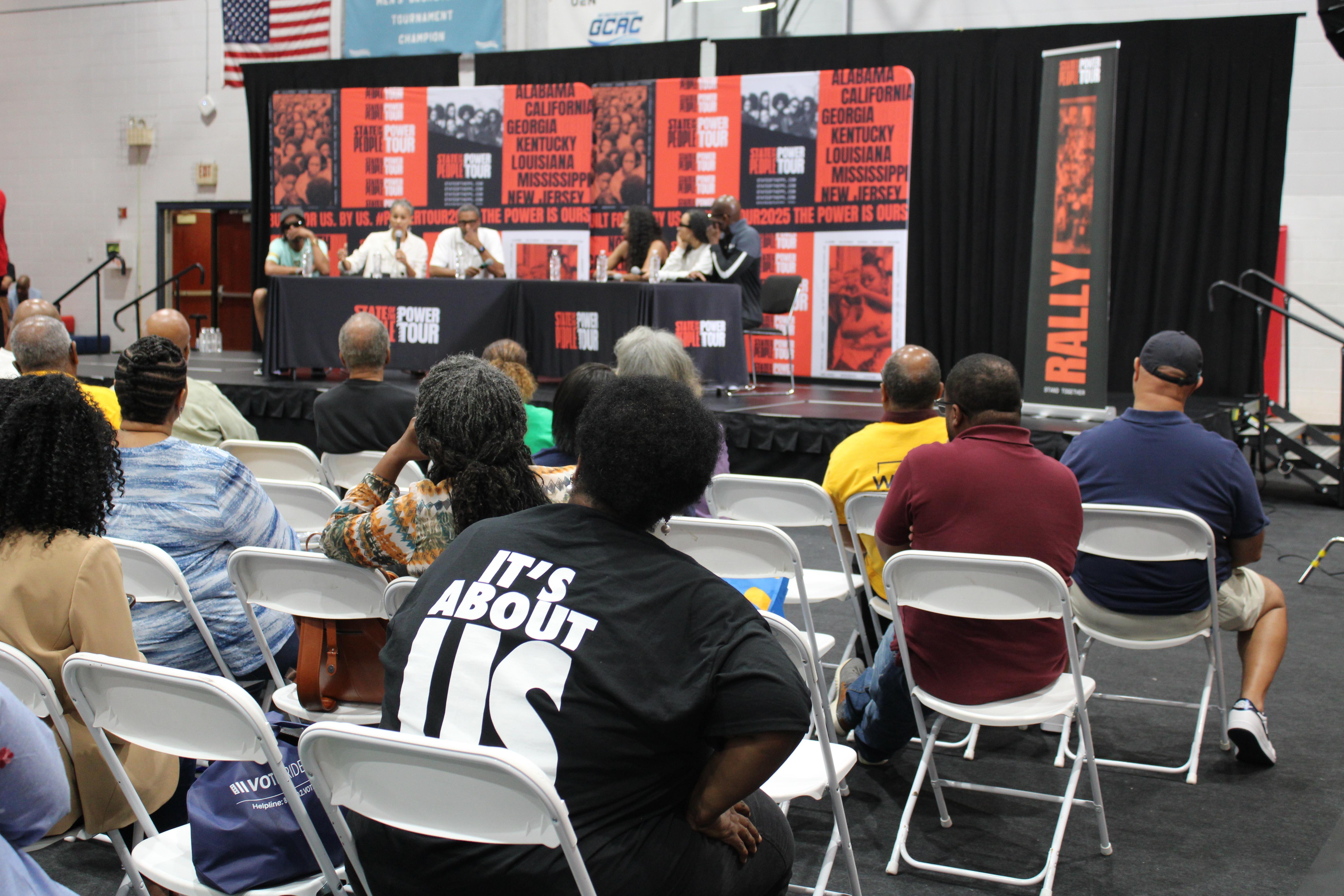 A woman wearing a black t-shirt with 'It's about us' printed on the back, listens to a panel.