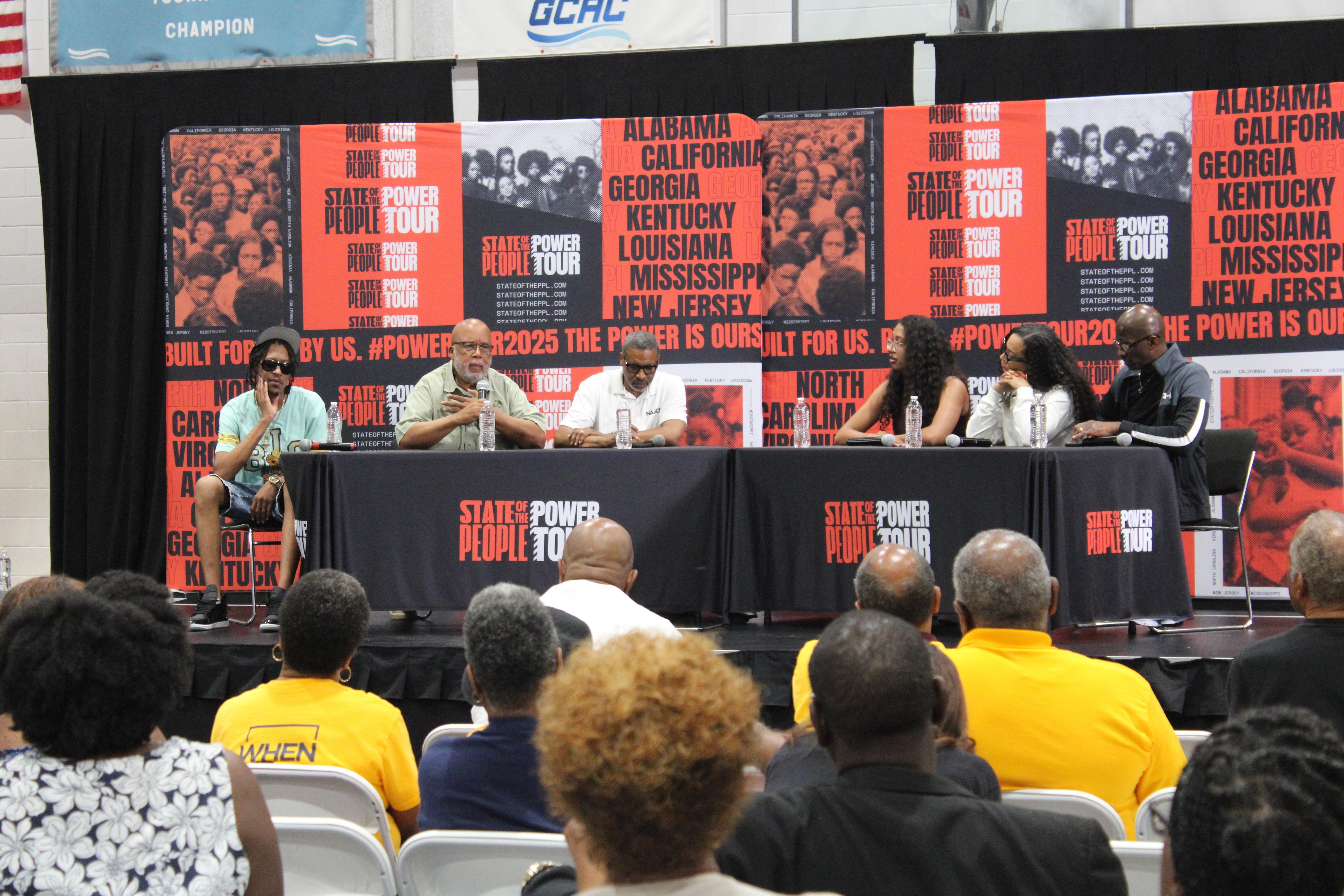 A man holds a microphone while sitting on a panel in front of an audience.