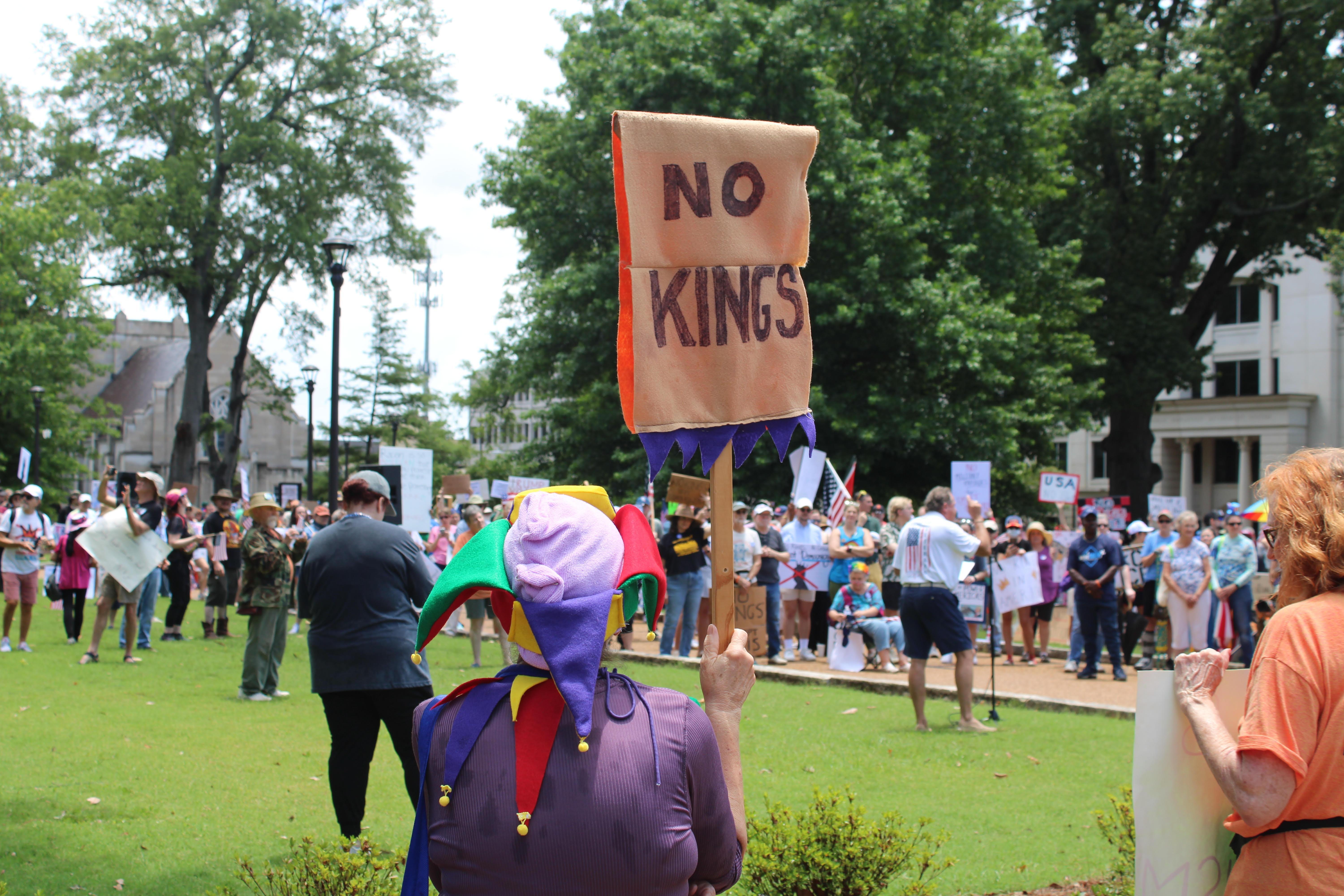 A woman wears a multicolor jester's hat while holding a sign reading 'No Kings.'