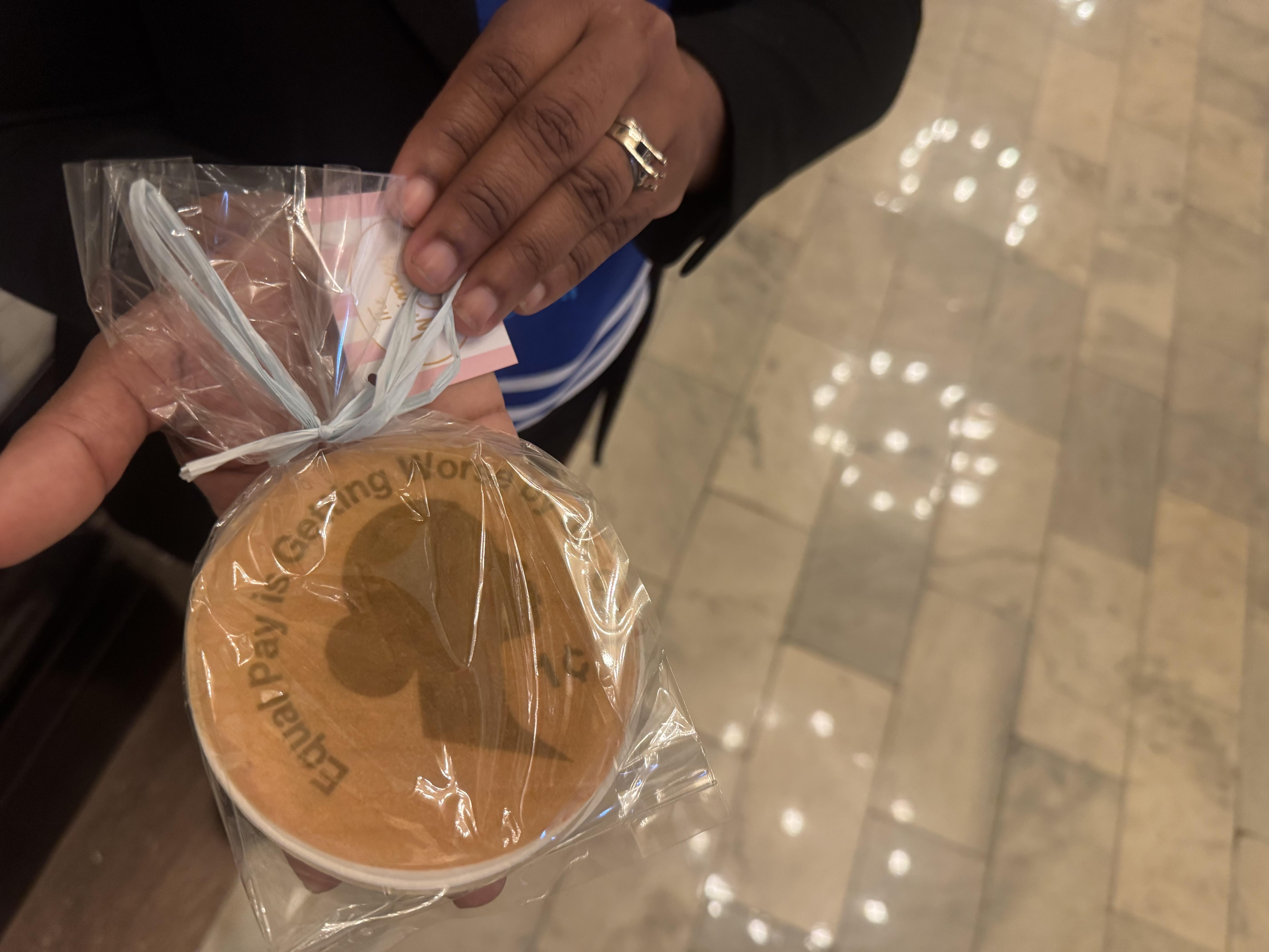 A woman holds a cookie with the outline of a woman and the words equal pay is getting worse by the cent.