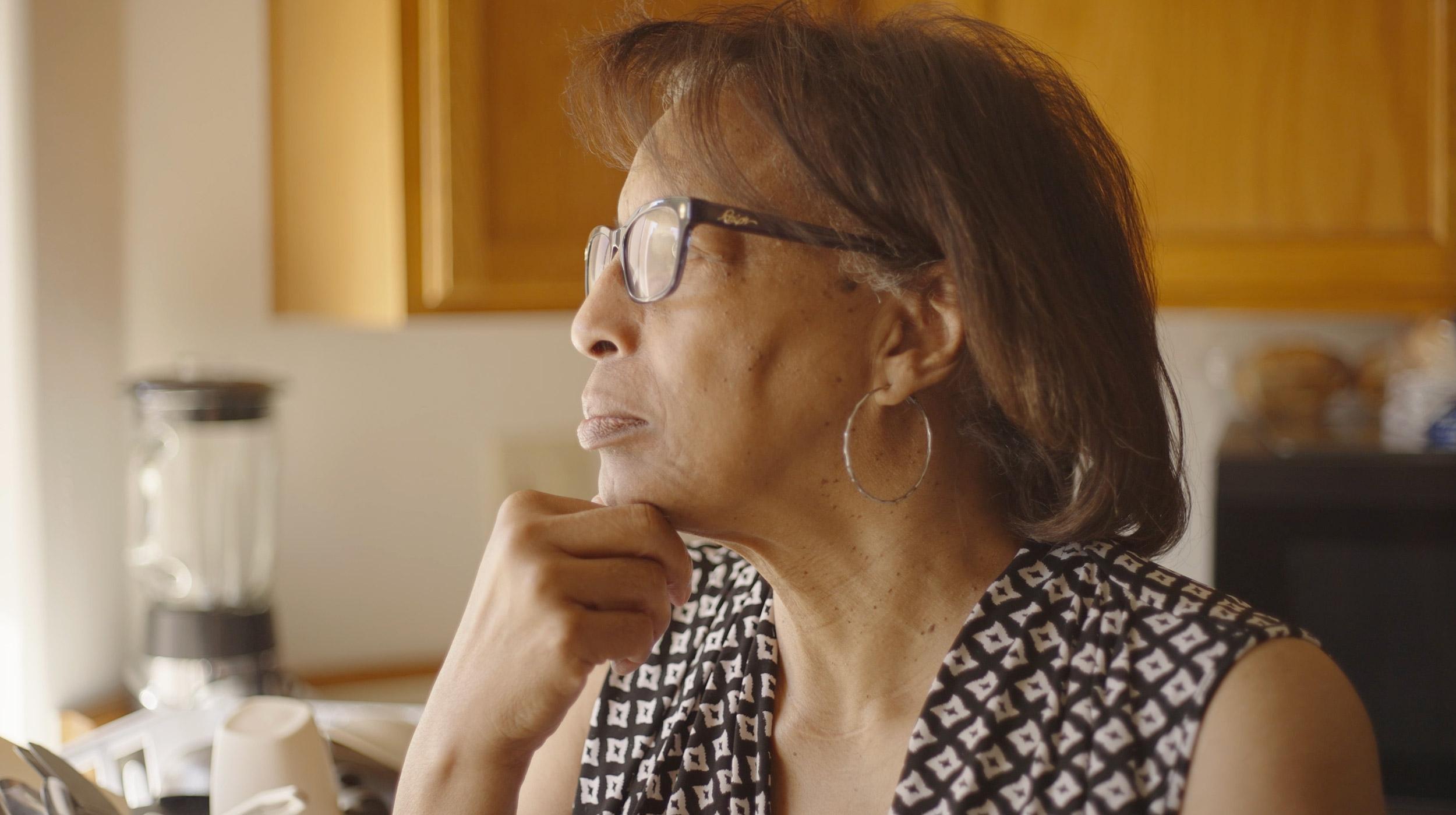 A photo shows an older Black woman with straight, shoulder-length brown hair, hoop earrings and glasses looking to the side with her hand under her chin. 