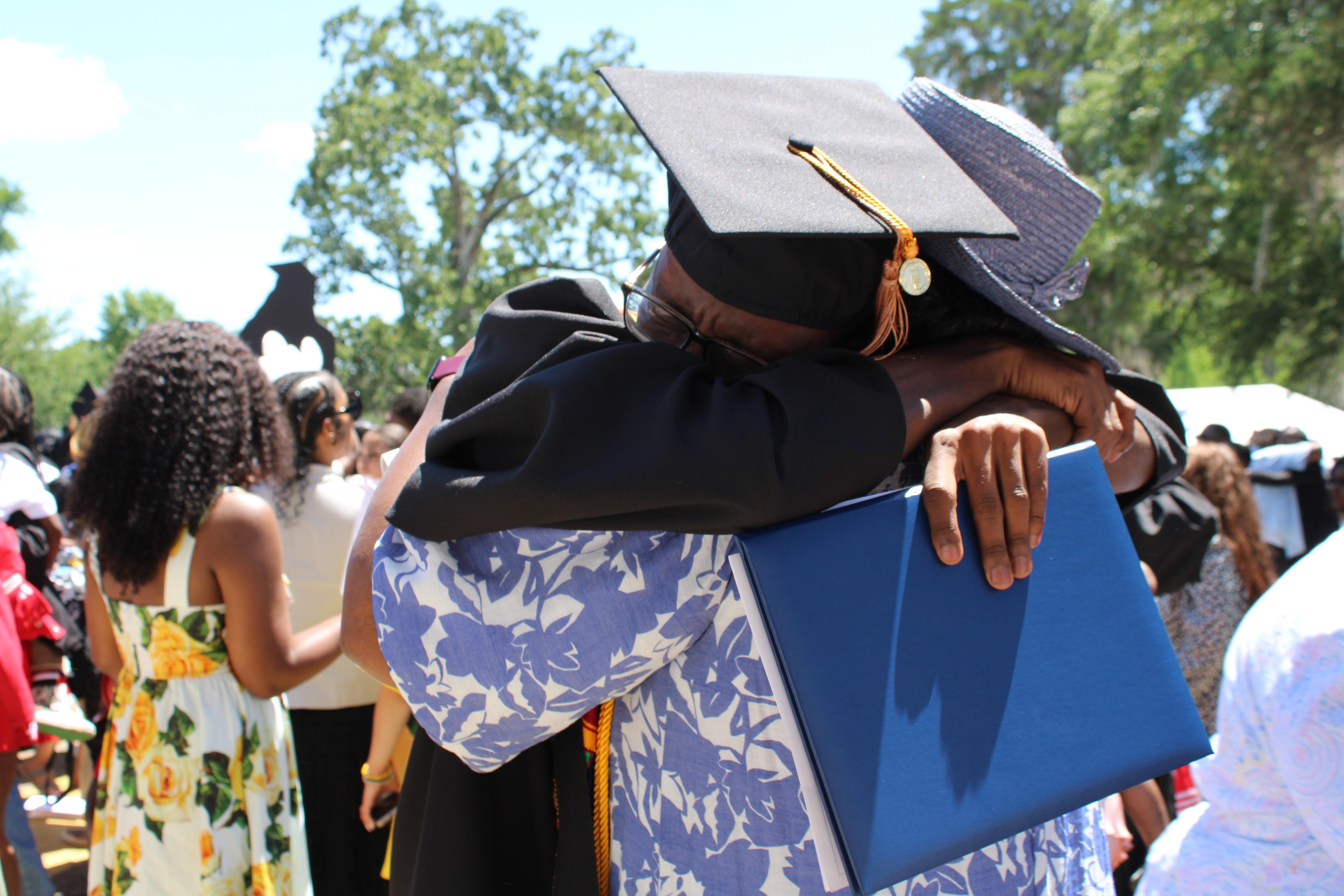 A young graduate tearfully hugs his grandmother while holding his diploma.